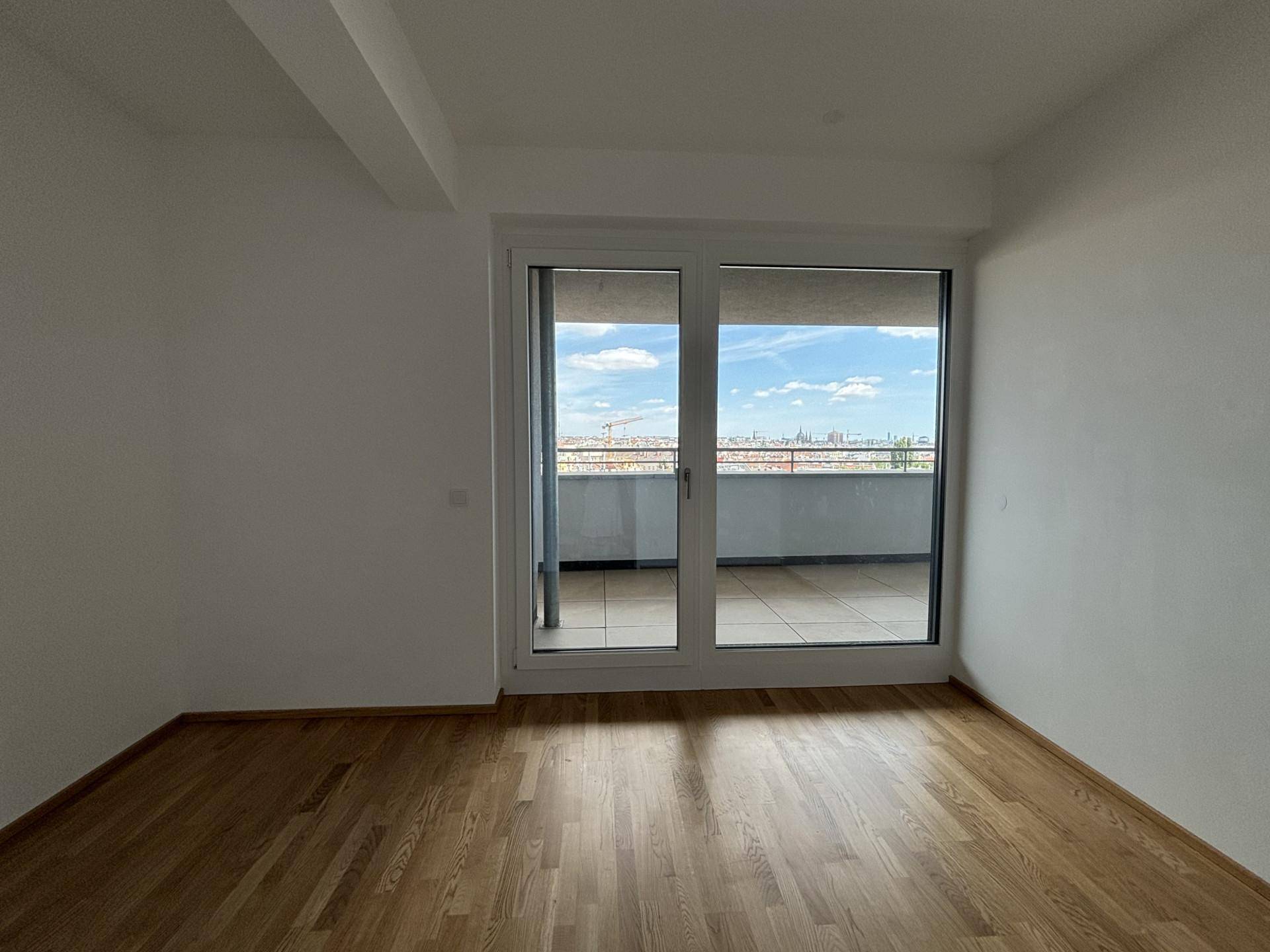 Leeres Zimmer mit Holzboden und großen Fenstern, die Zugang zu einem Balkon bieten.