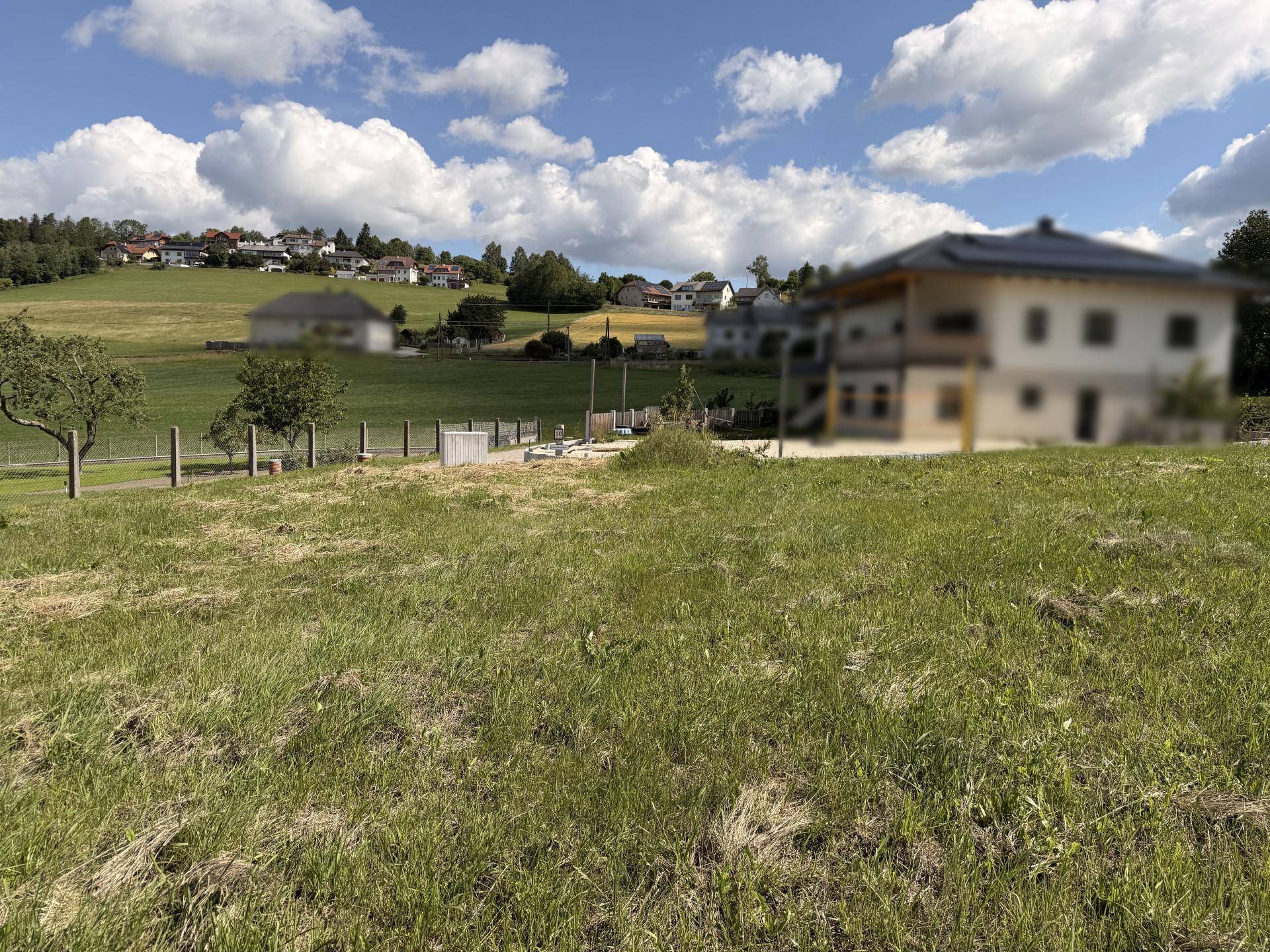 Grüne Wiese mit Blick auf ein Dorf am Hang und ein unscharfes Gebäude im Vordergrund.