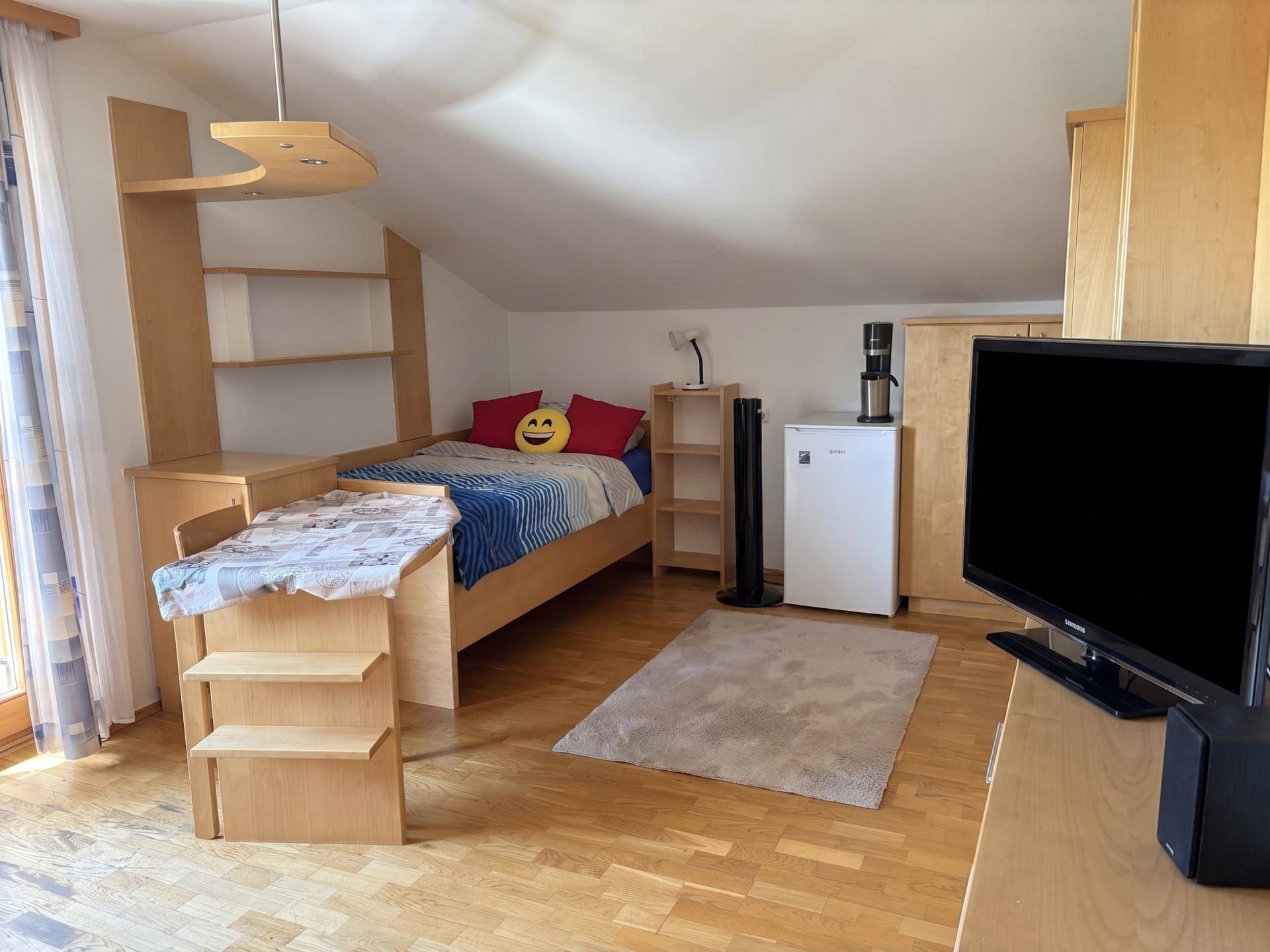 Helles Schlafzimmer mit Holzboden, Bett, Regalen und Fernseher im Dachgeschoss.