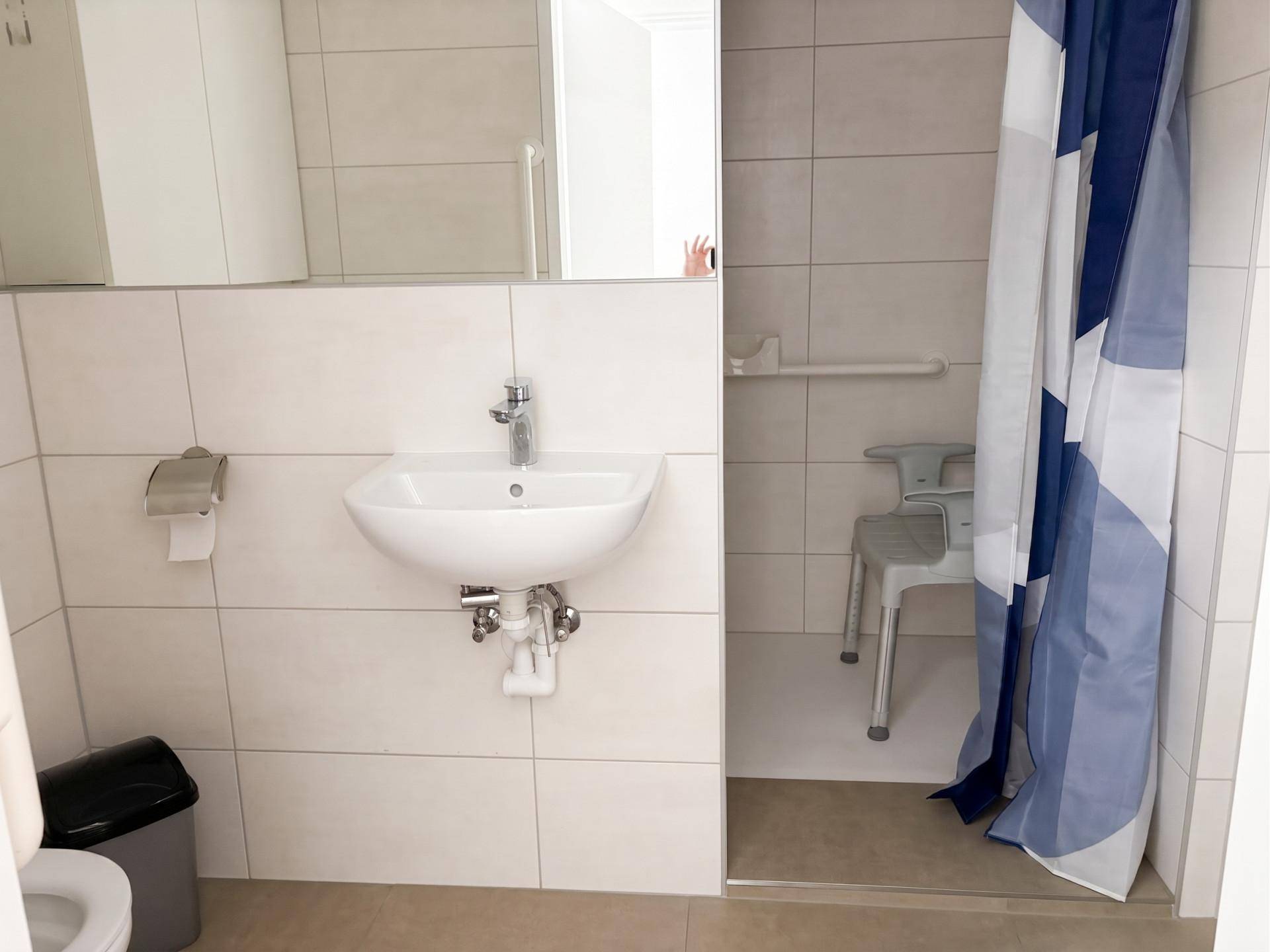 Barrierefreie Dusche mit Haltegriffen und Duschstuhl in einem hell gefliesten Badezimmer.