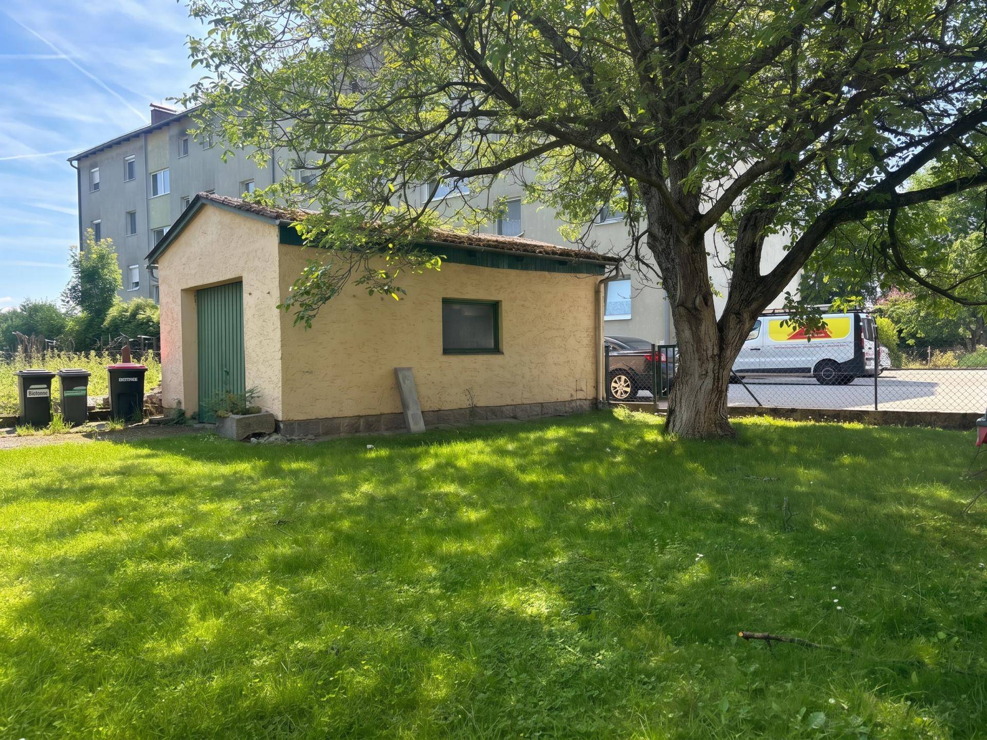 Kleiner Schuppen im Gartenbereich vor einem Mehrfamilienhaus, umgeben von Grün.