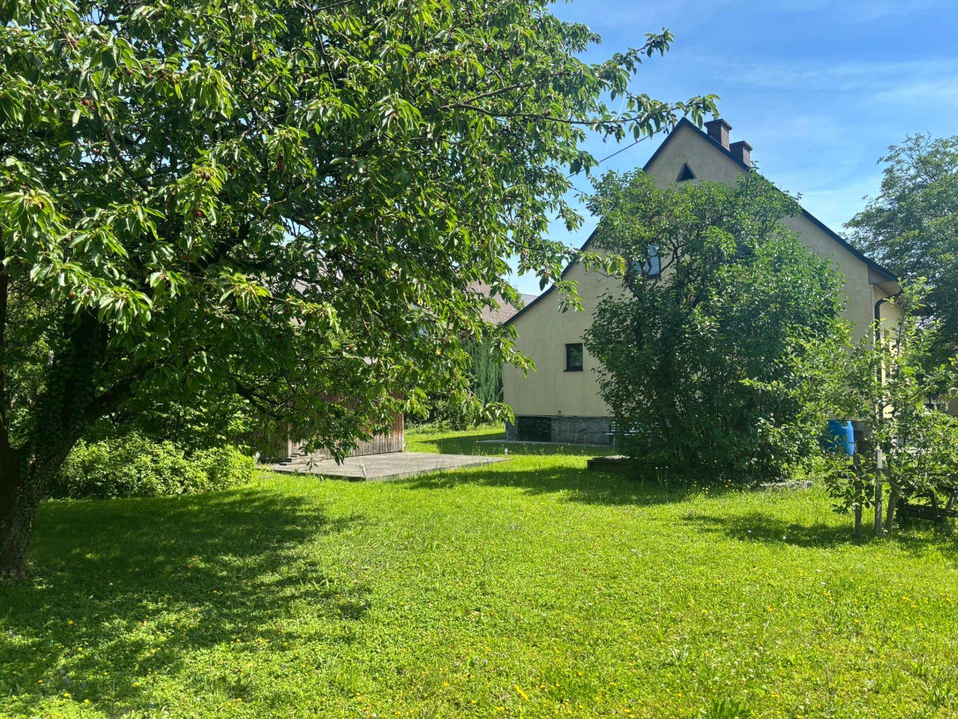 Seitenansicht des Hauses mit großem Garten und altem Baumbestand unter blauem Himmel.