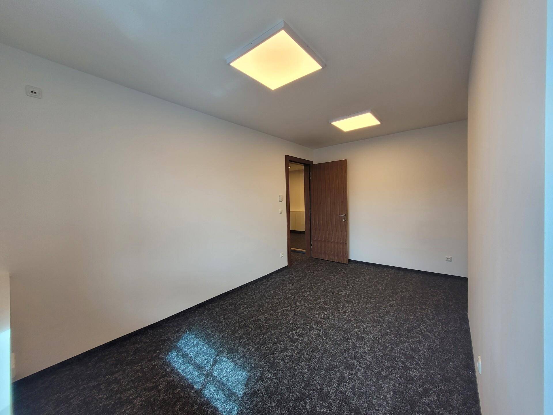 Leerer Büroraum mit Teppichboden und einer Holztür, die zu einem weiteren Bereich führt.