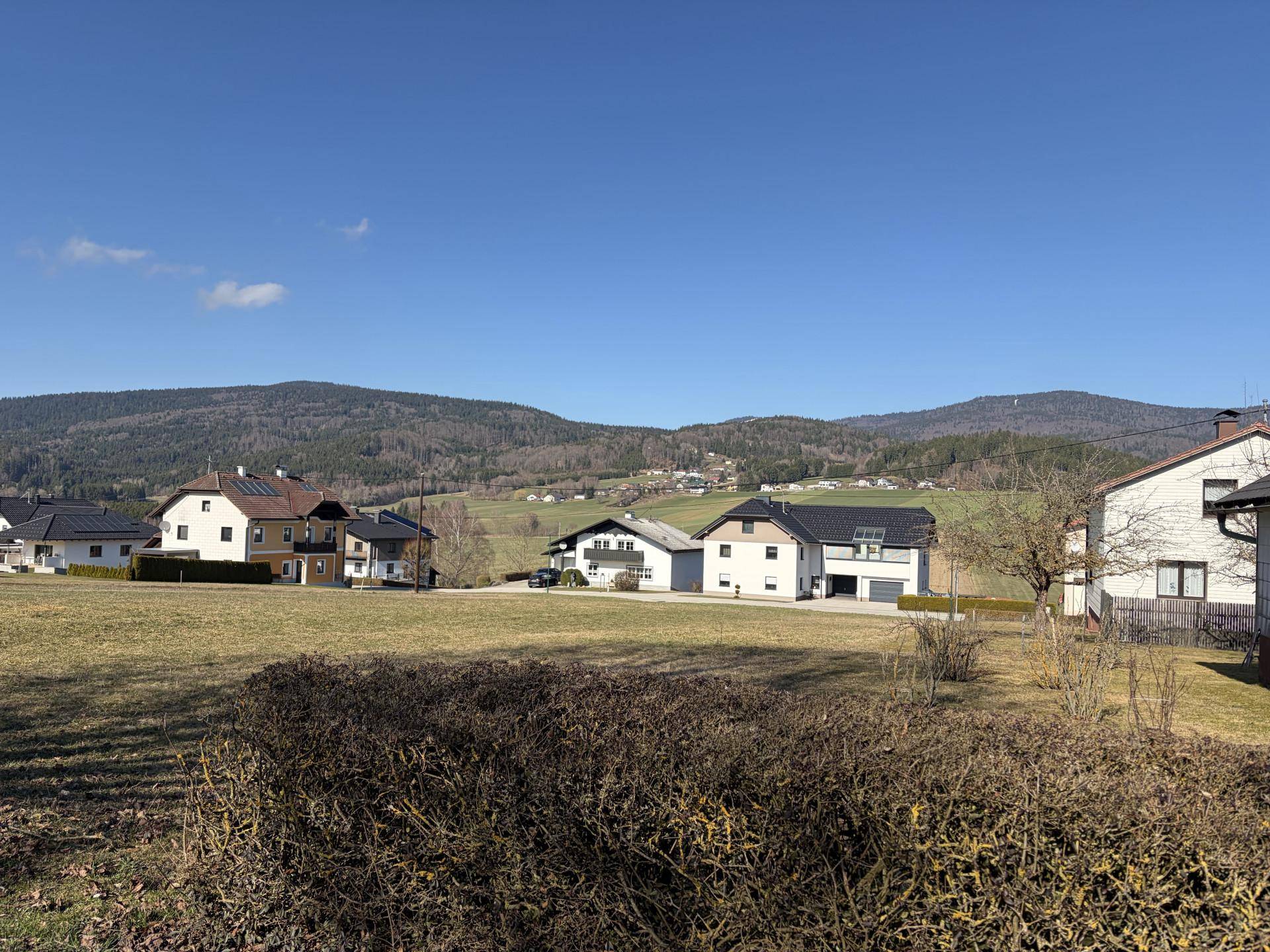Wohngebiet mit modernen Häusern, umgeben von grünen Wiesen und bewaldeten Bergen unter einem strahlend blauen Himmel.