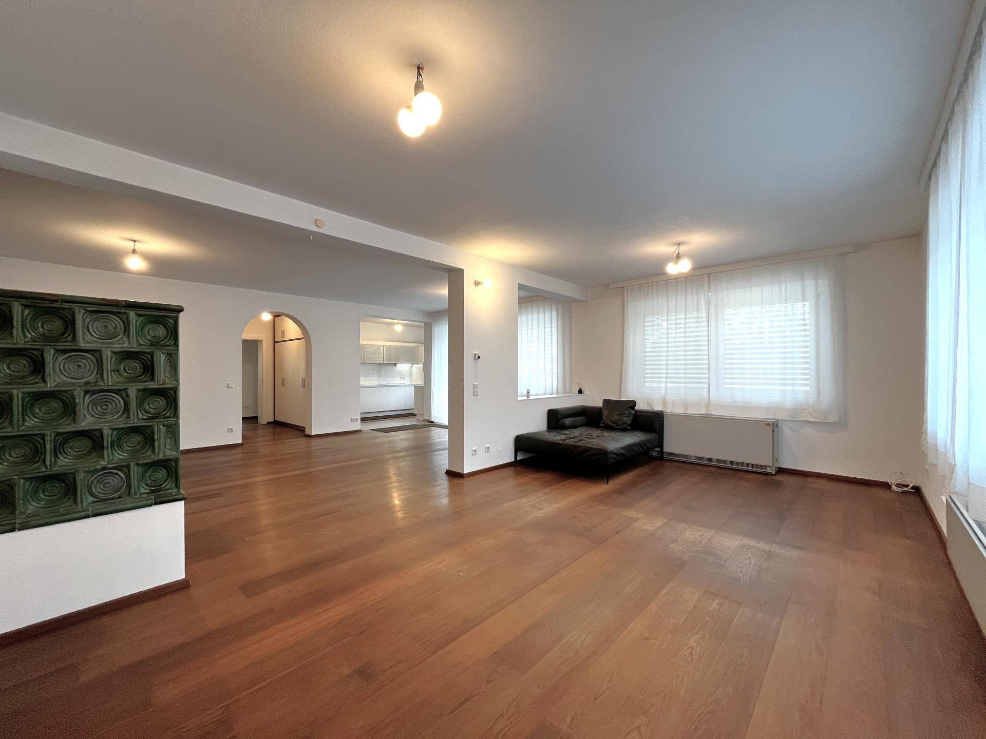 Geräumiges Wohnzimmer mit dunklem Parkettboden, Kachelofen und Blick in die Küche.