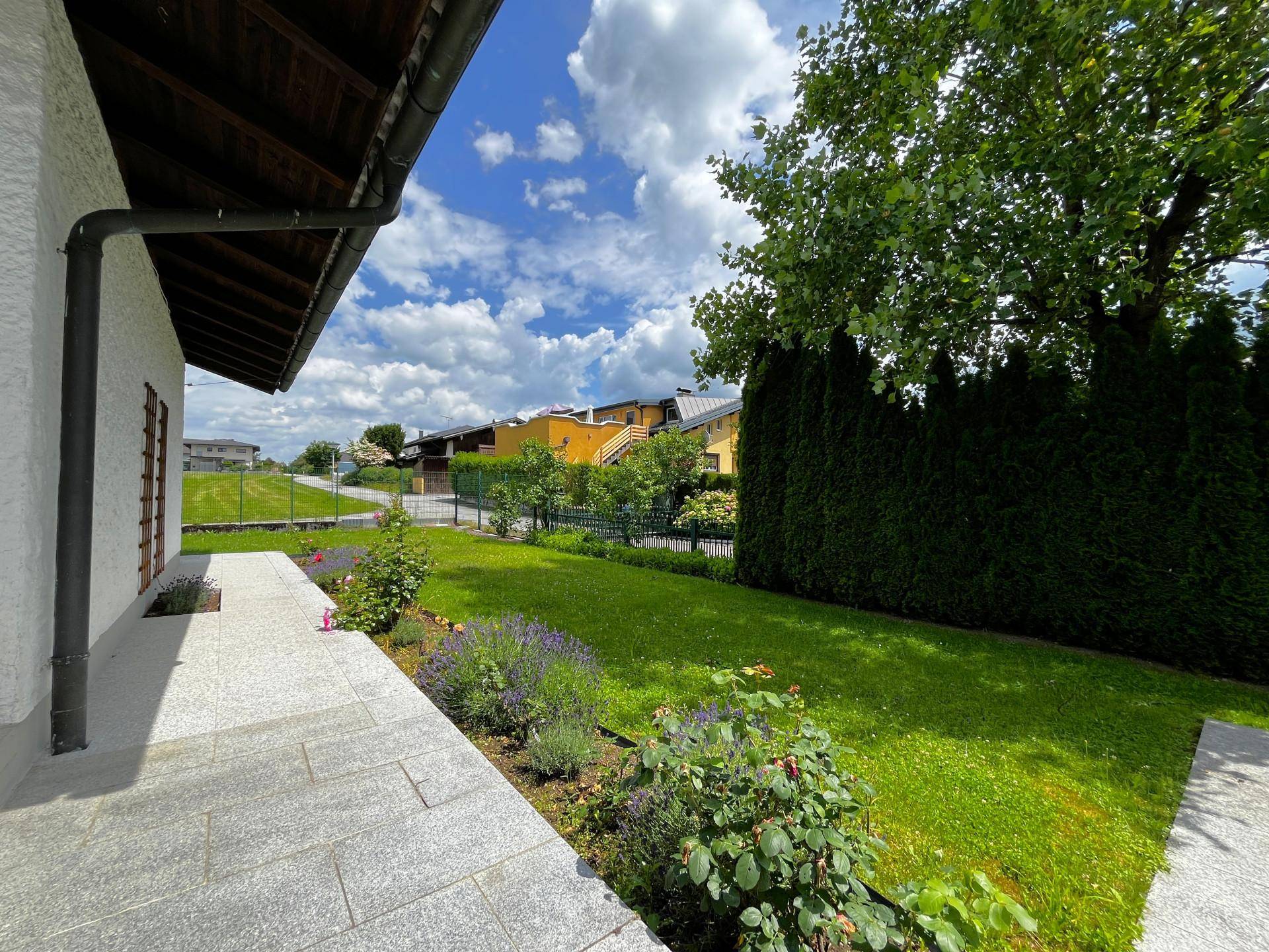 Gepflegter Gartenbereich mit Rasenfläche, Blumenbeeten und Hecke unter blauem Himmel.
