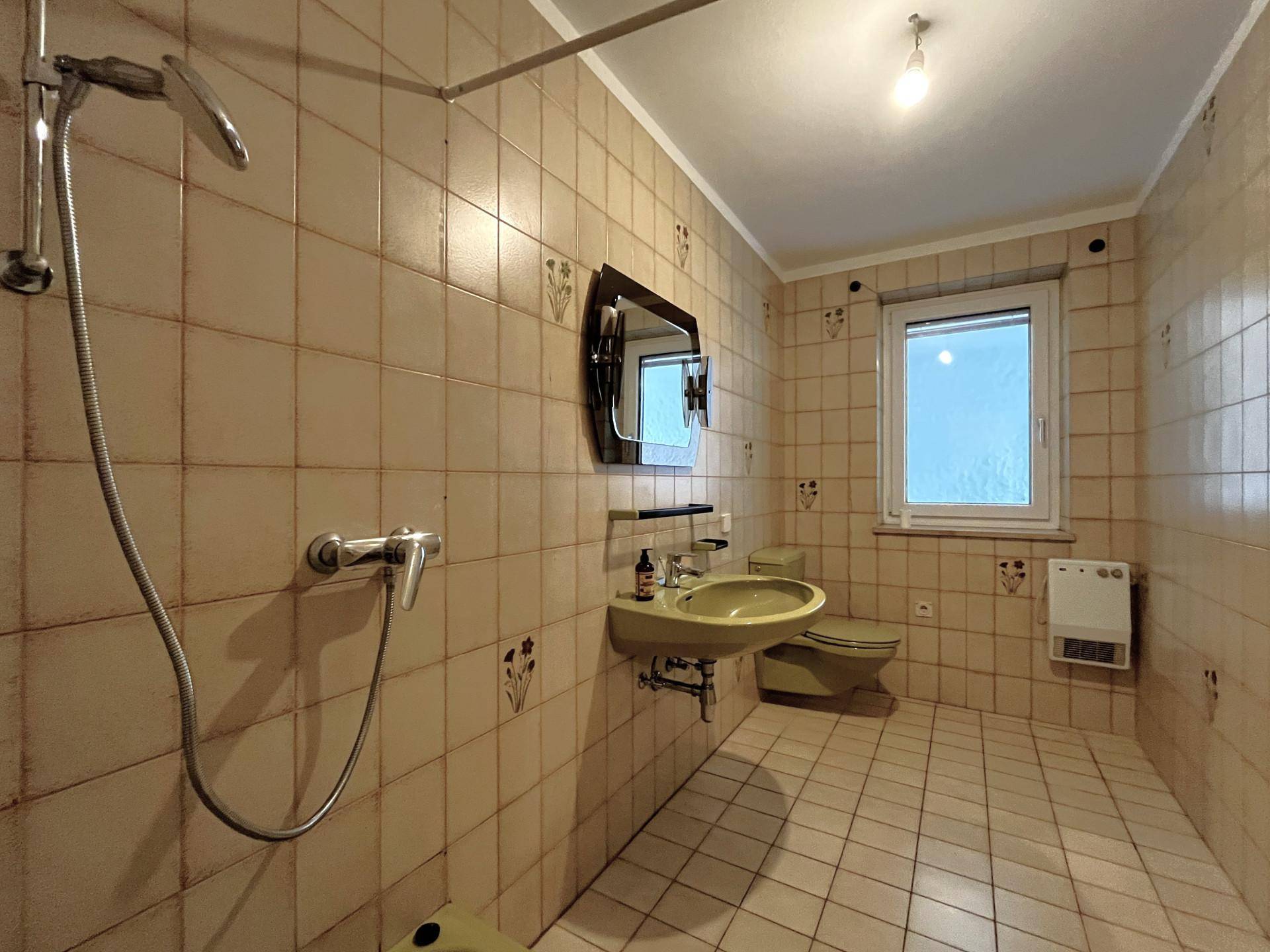 Badezimmer mit beigefarbenen Fliesen, Dusche und Fenster, das Tageslicht hereinlässt.