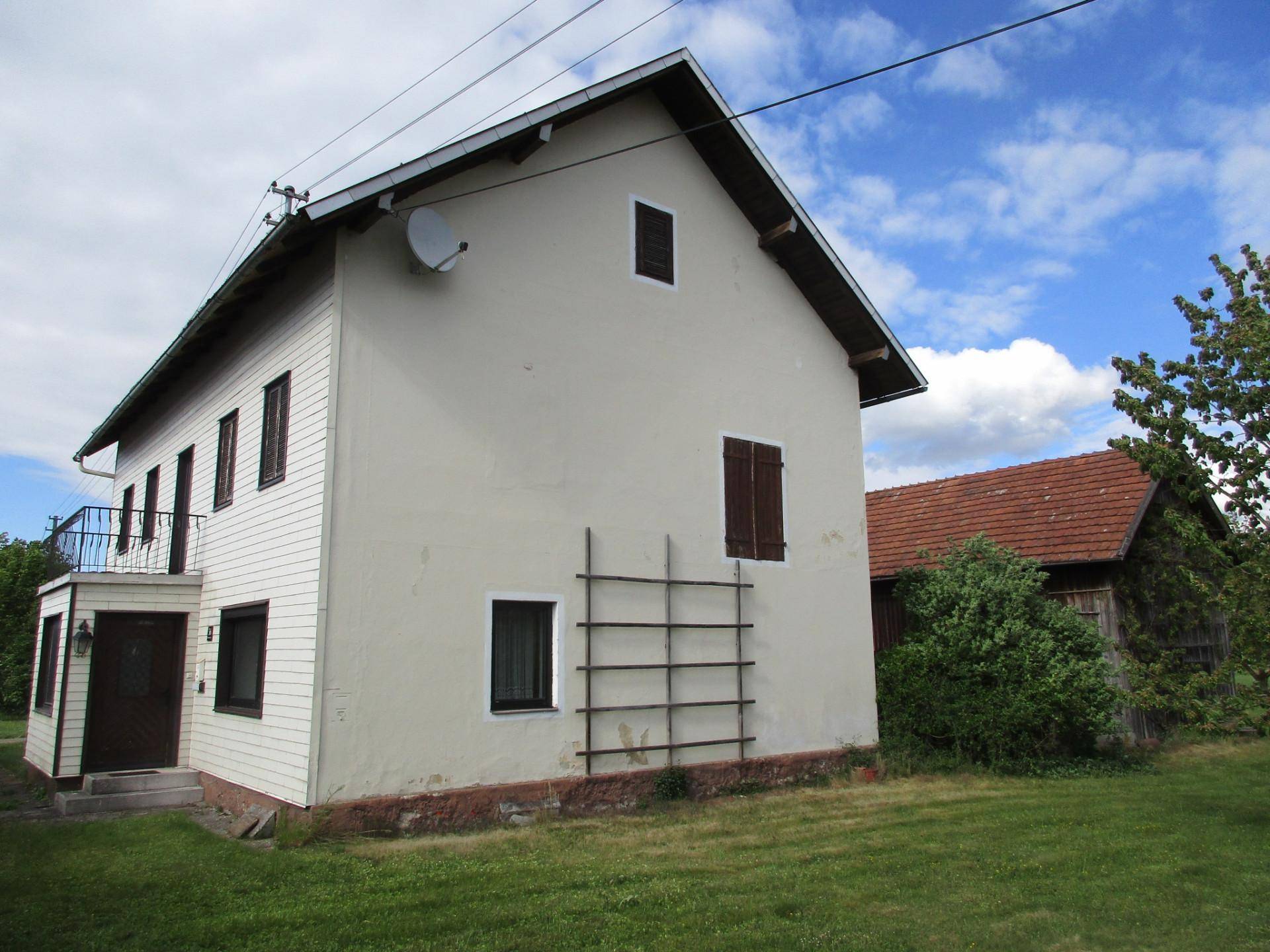 Ländliches Haus mit heller Fassade, Holzverkleidung und einem kleinen Balkon unter blauem Himmel.