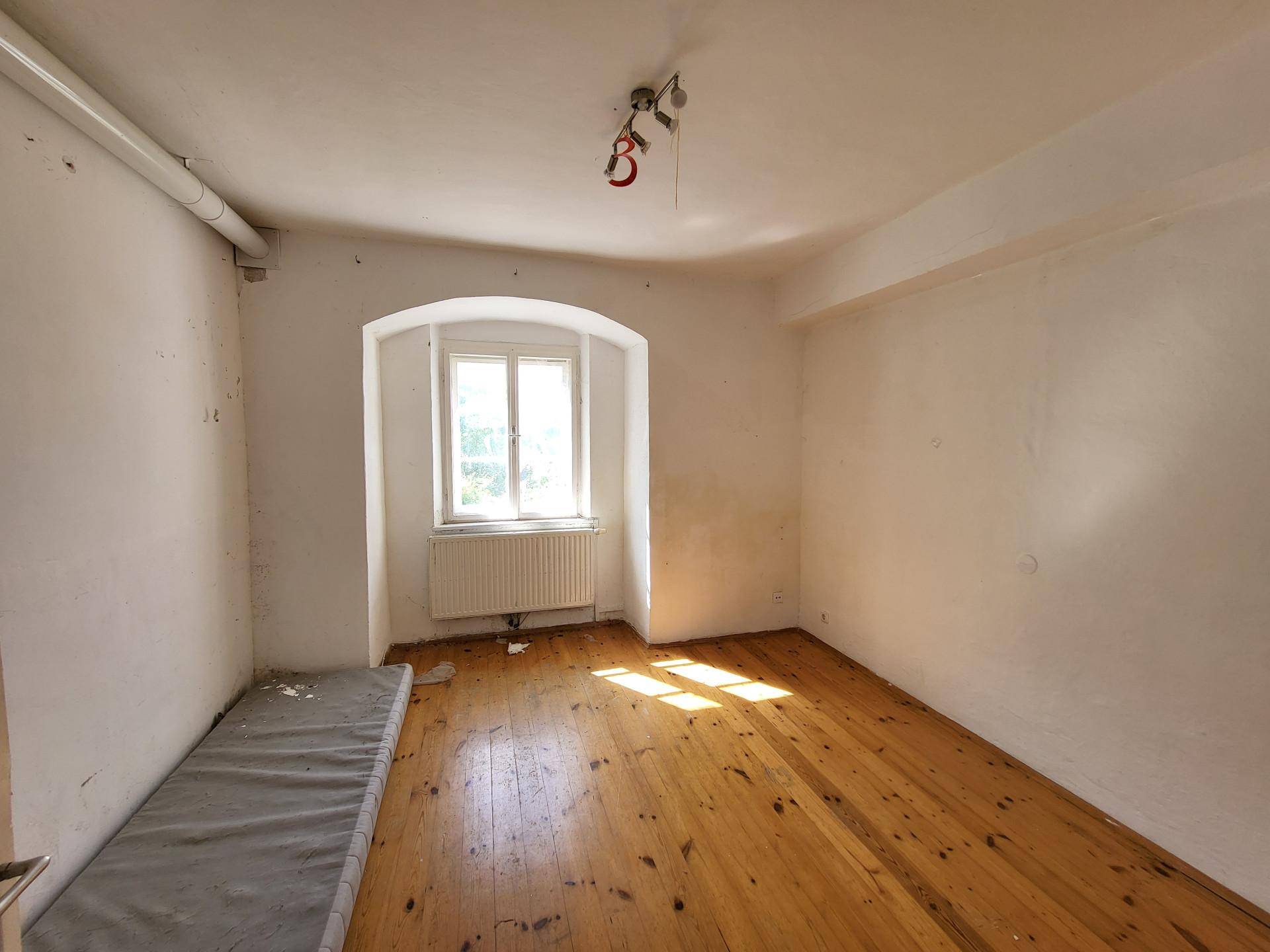 Helles Zimmer mit Holzdielenboden, einem Fenster und einem Heizkörper, bereit zur Renovierung.