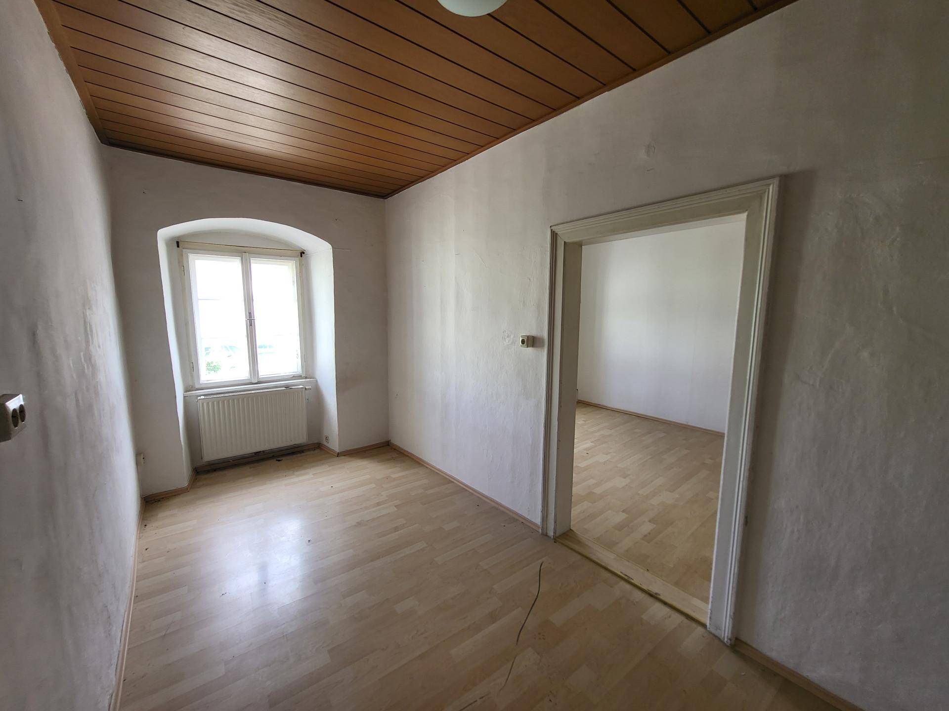 Kleines Zimmer mit Holzdecke, Laminatboden und einem Fenster, das in einen weiteren Raum führt.