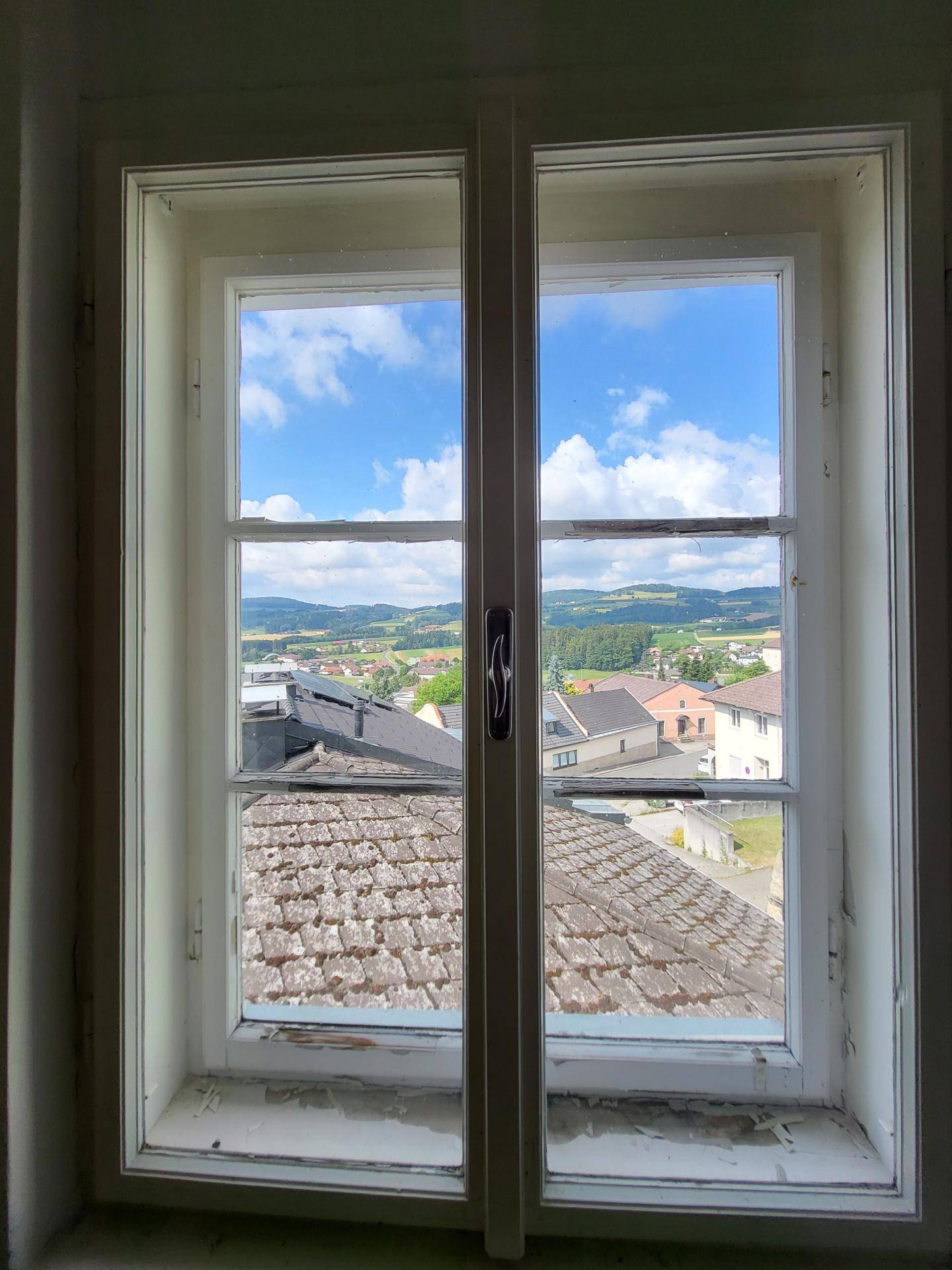 Fensterblick auf die umliegende Landschaft mit Häusern und grünen Hügeln unter blauem Himmel.