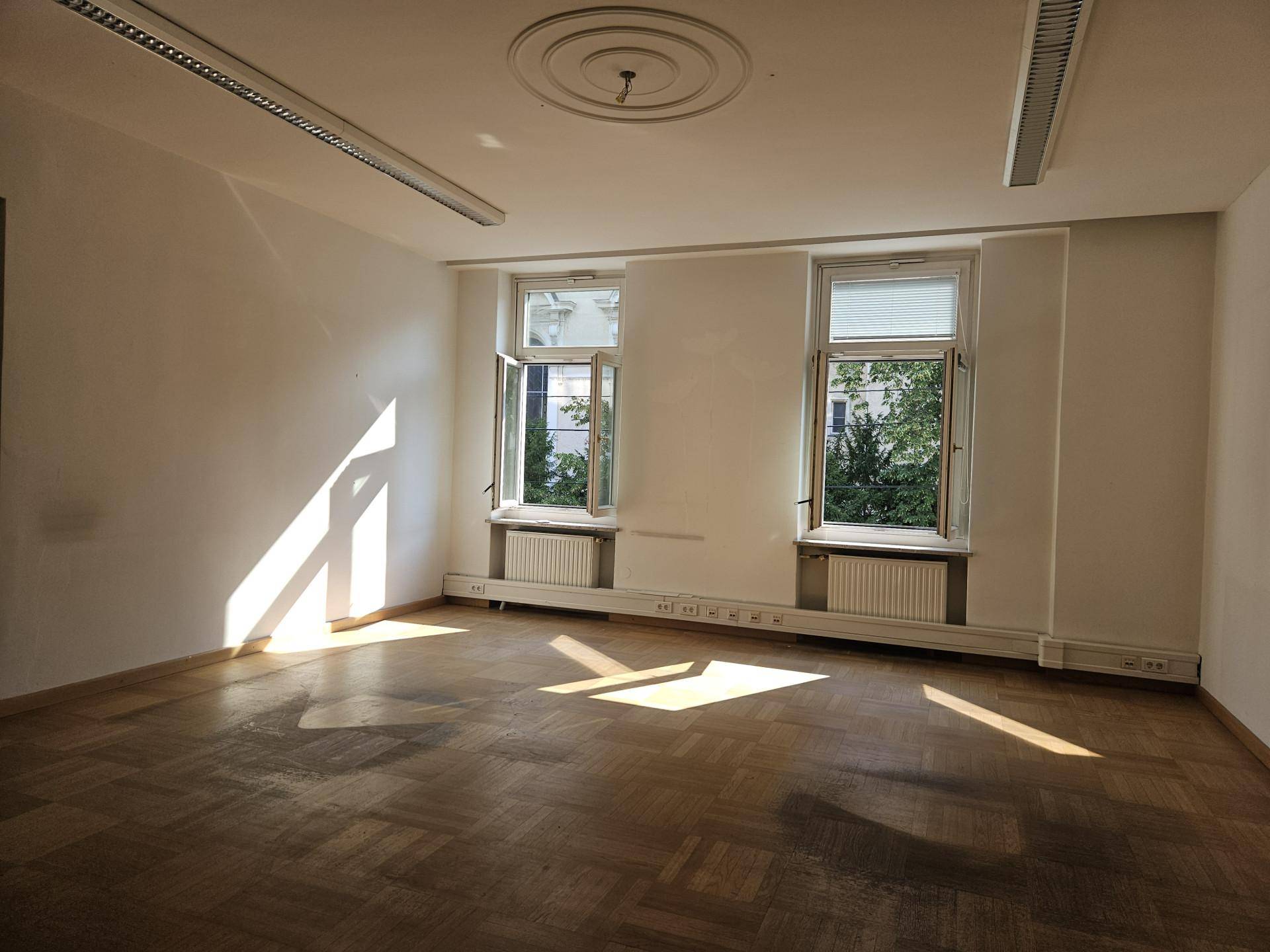 Heller, leerer Raum mit Parkettboden, zwei Fenstern und einem klassischen Deckenstuck.