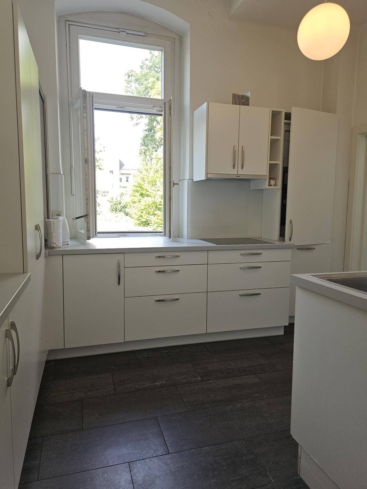 Moderne weiße Küche mit dunklem Fliesenboden und einem großen Fenster, das viel Licht hereinlässt.