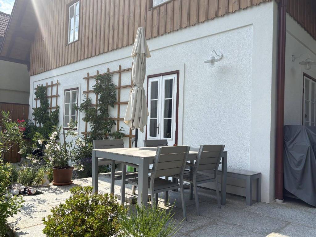 Sonniger Außenbereich mit Esstisch, Stühlen und Sonnenschirm, umgeben von Pflanzen.