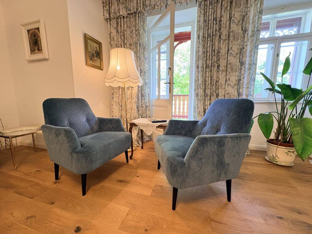 Gemütliches Wohnzimmer mit zwei blauen Samtsesseln, Holzboden und Zugang zu einem Balkon.