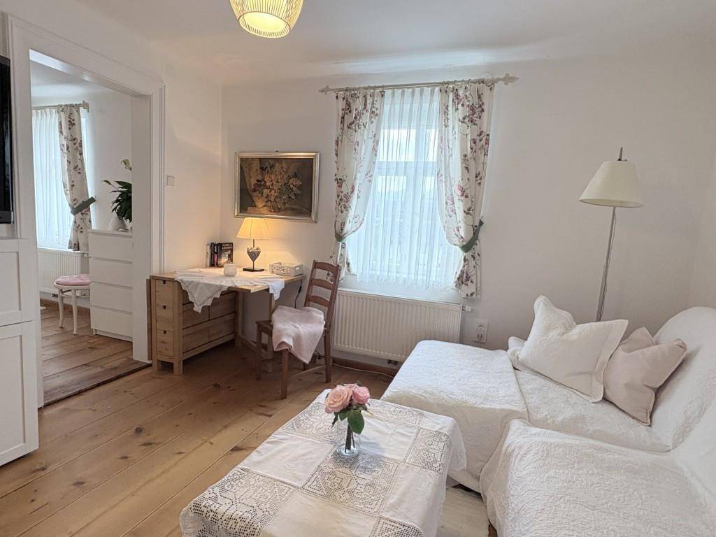 Gemütliches Wohnzimmer mit weißem Sofa, Schreibtisch und floralen Vorhängen, auf Holzboden.