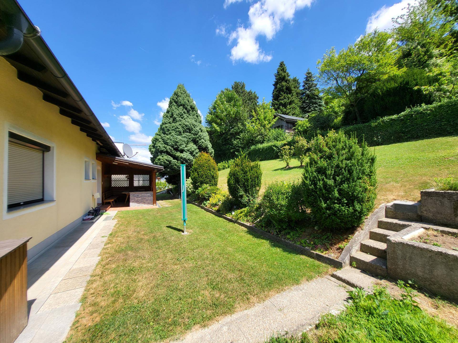 Seitenansicht des Hauses mit überdachtem Außenbereich und dem terrassierten Garten in Hanglage.