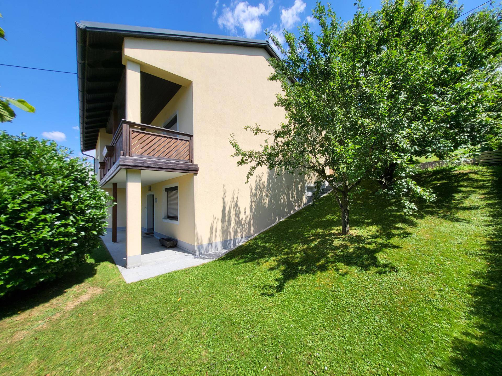 Eckansicht des Hauses mit Balkon und Holzgeländer, eingebettet in die grüne Hanglandschaft.