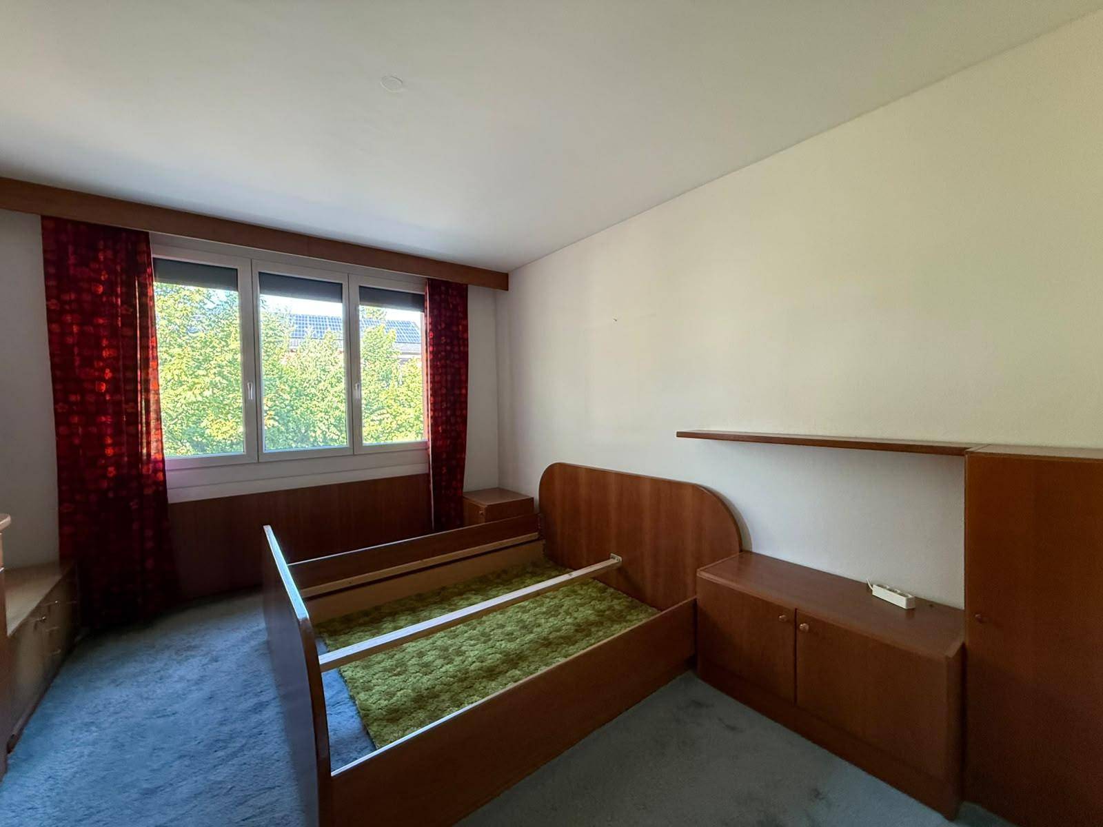 Schlafzimmer mit Teppichboden, Bettrahmen und Fenster mit Blick auf Bäume.