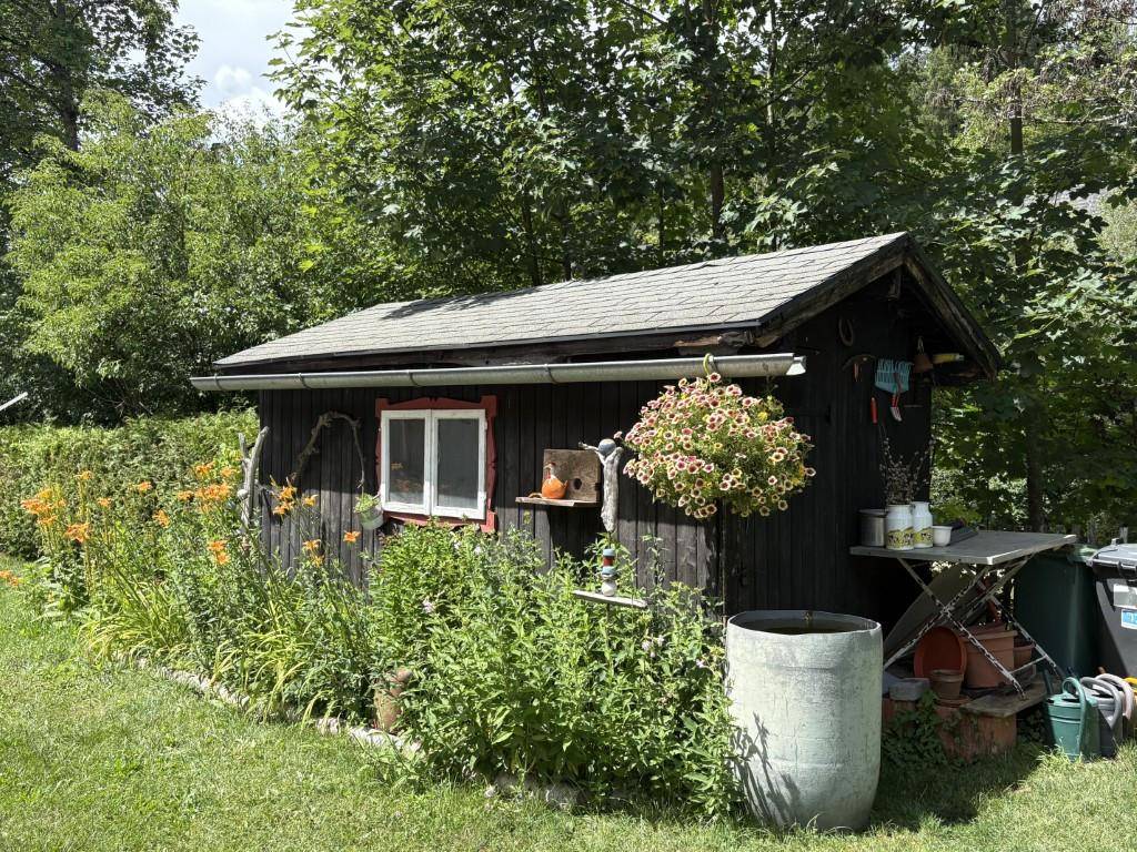 Kleiner Holzschuppen im Garten, umgeben von Blumen und Pflanzen.