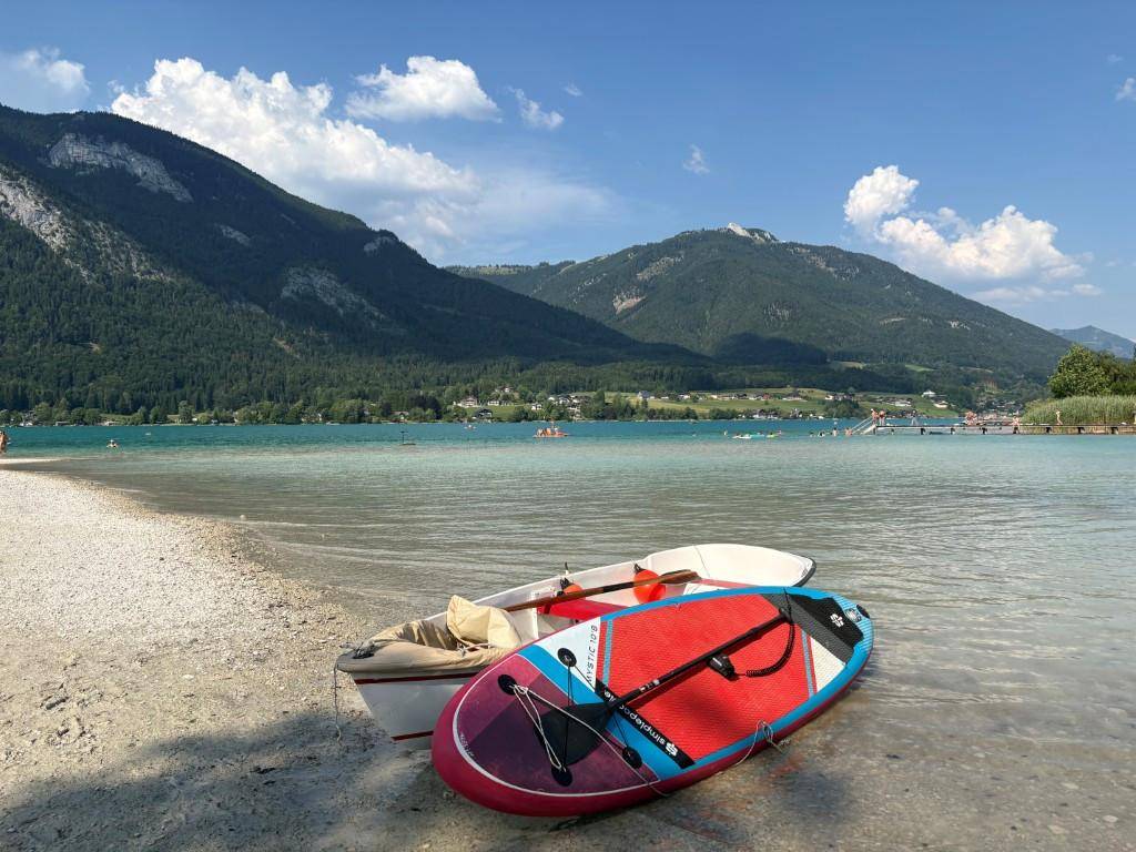 Kiesstrand am See mit Booten und Blick auf die umliegenden Berge.