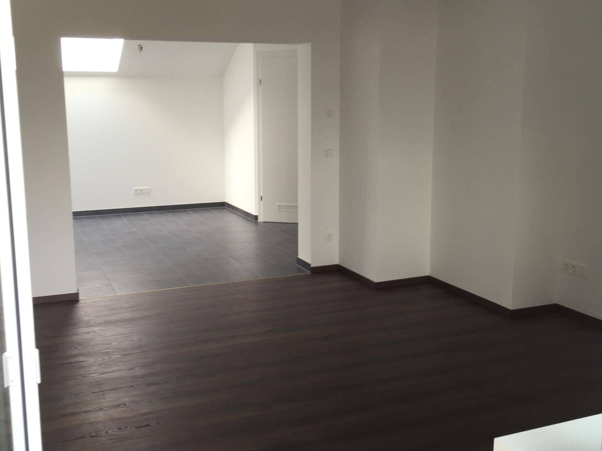Helle, leere Räume mit dunklem Holzboden und Fliesen, verbunden durch einen offenen Durchgang und Oberlicht.