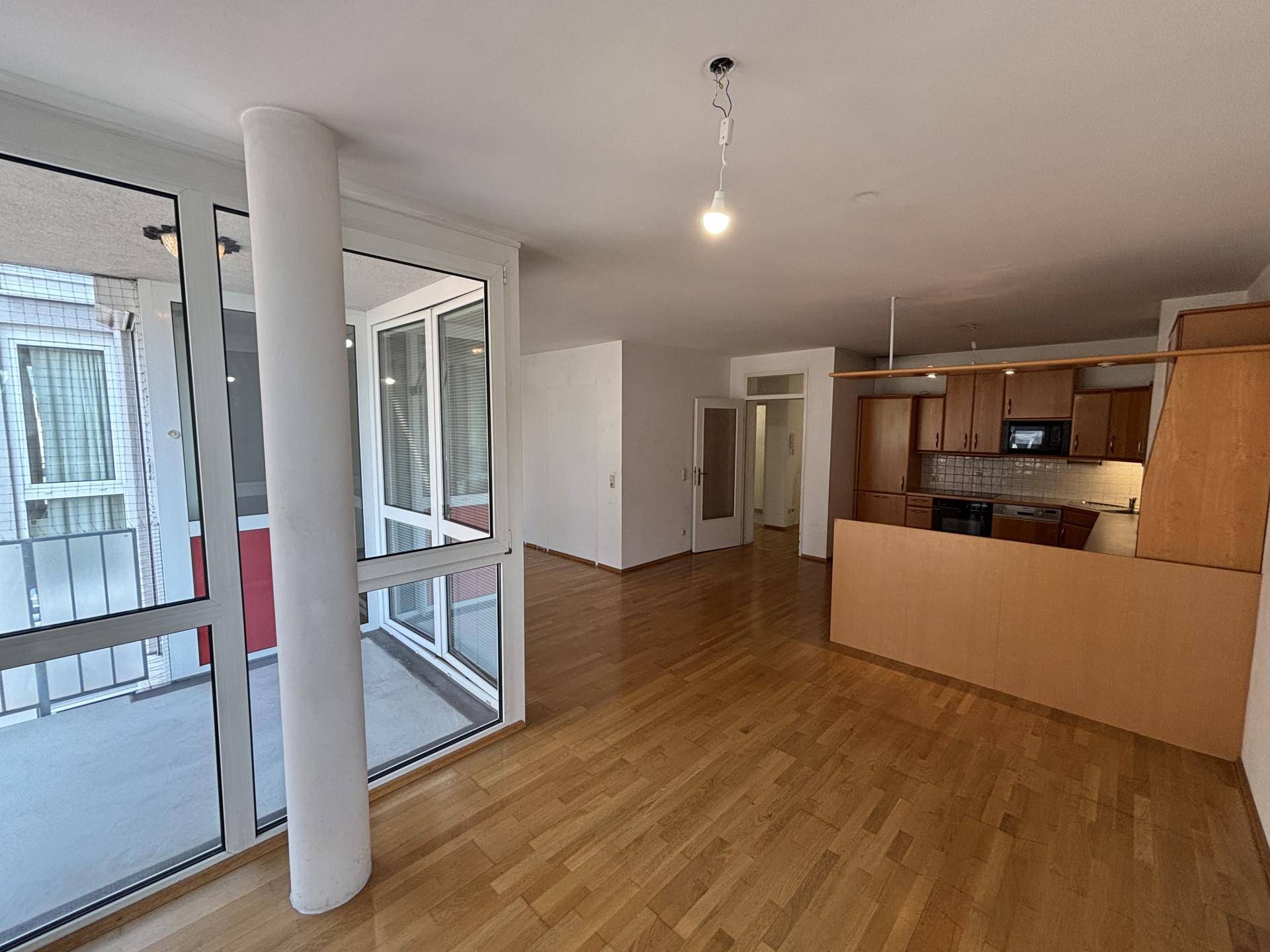 Lichtdurchfluteter Wohnbereich mit Holzboden, Zugang zu einem Balkon und offener Verbindung zur Küche.
