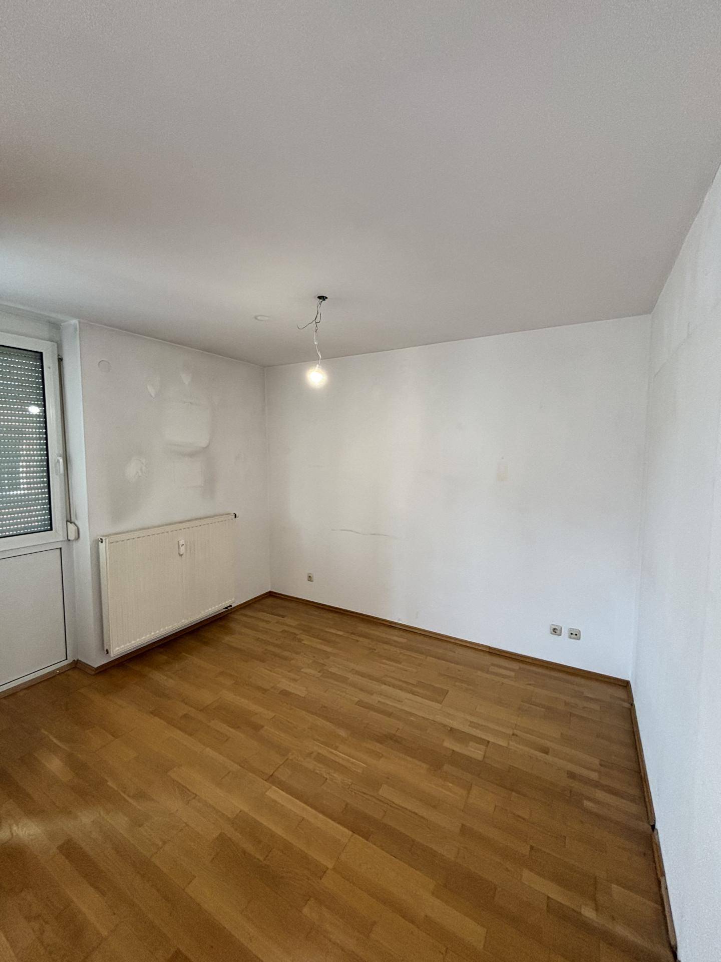 Leeres Zimmer mit Holzboden, weißen Wänden und einem Fenster mit Rollladen, bereit zur individuellen Gestaltung.