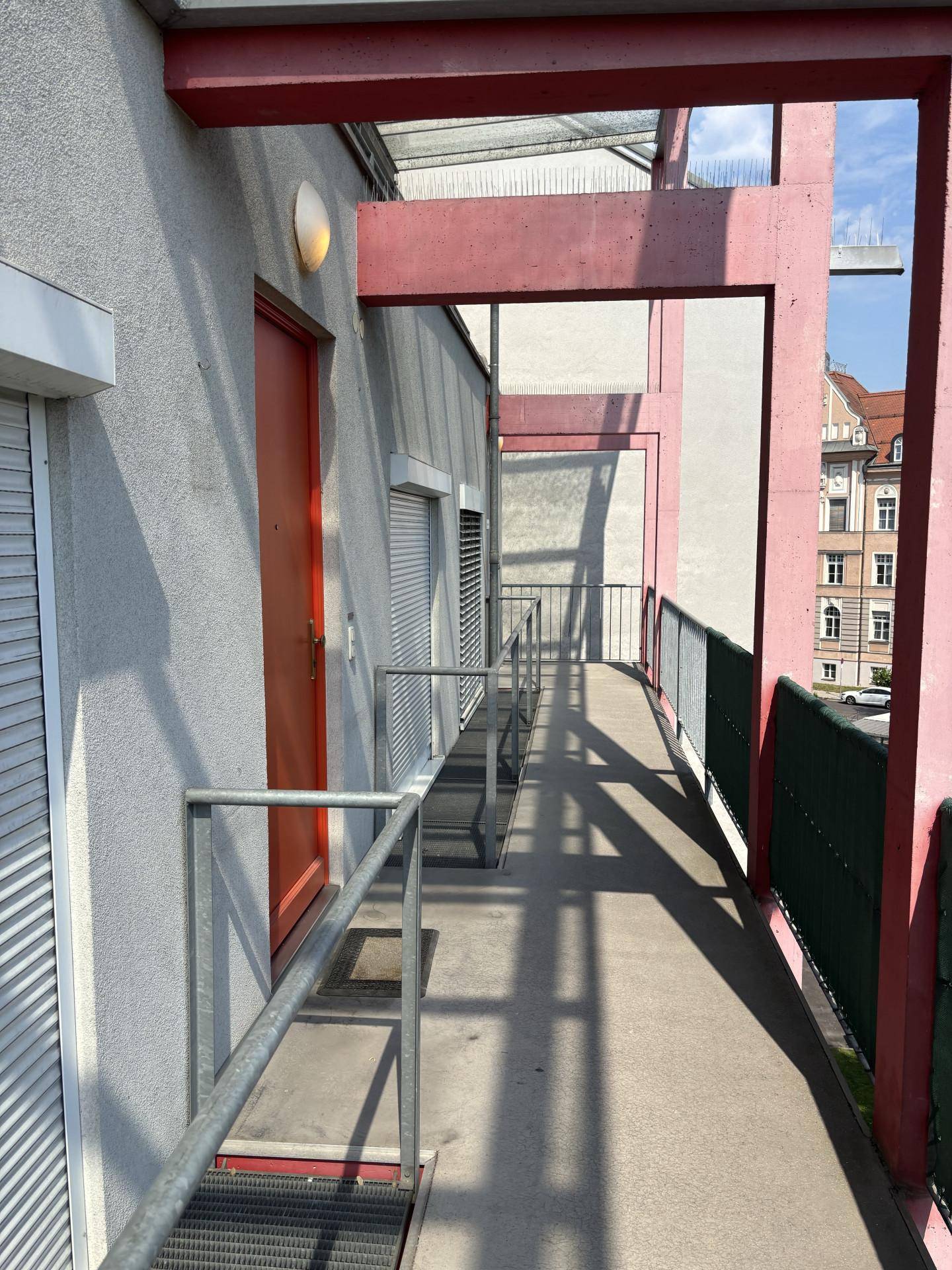 Langer Außenflur mit Rampe und Metallgeländer, der Zugang zu den Wohnungen bietet und von roten Trägern gestützt wird.