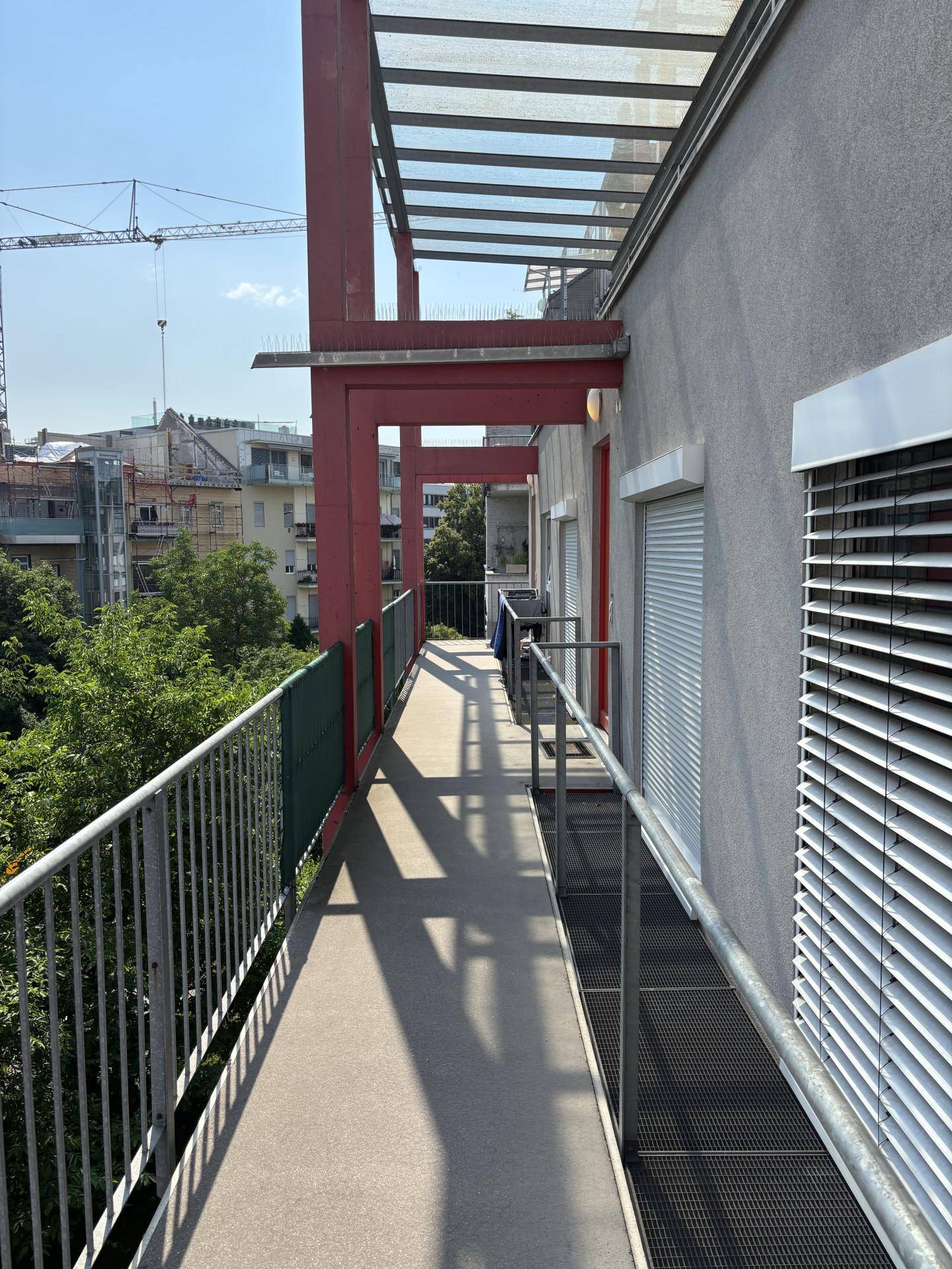Sonniger Außenbalkon mit Rampe und Blick auf Bäume und eine Baustelle in der Ferne.