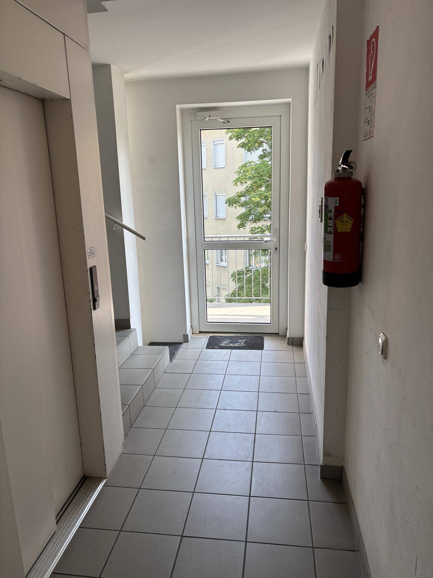 Heller Eingangsbereich mit Fliesenboden, Aufzug und einer Tür, die ins Freie führt, inklusive Feuerlöscher.