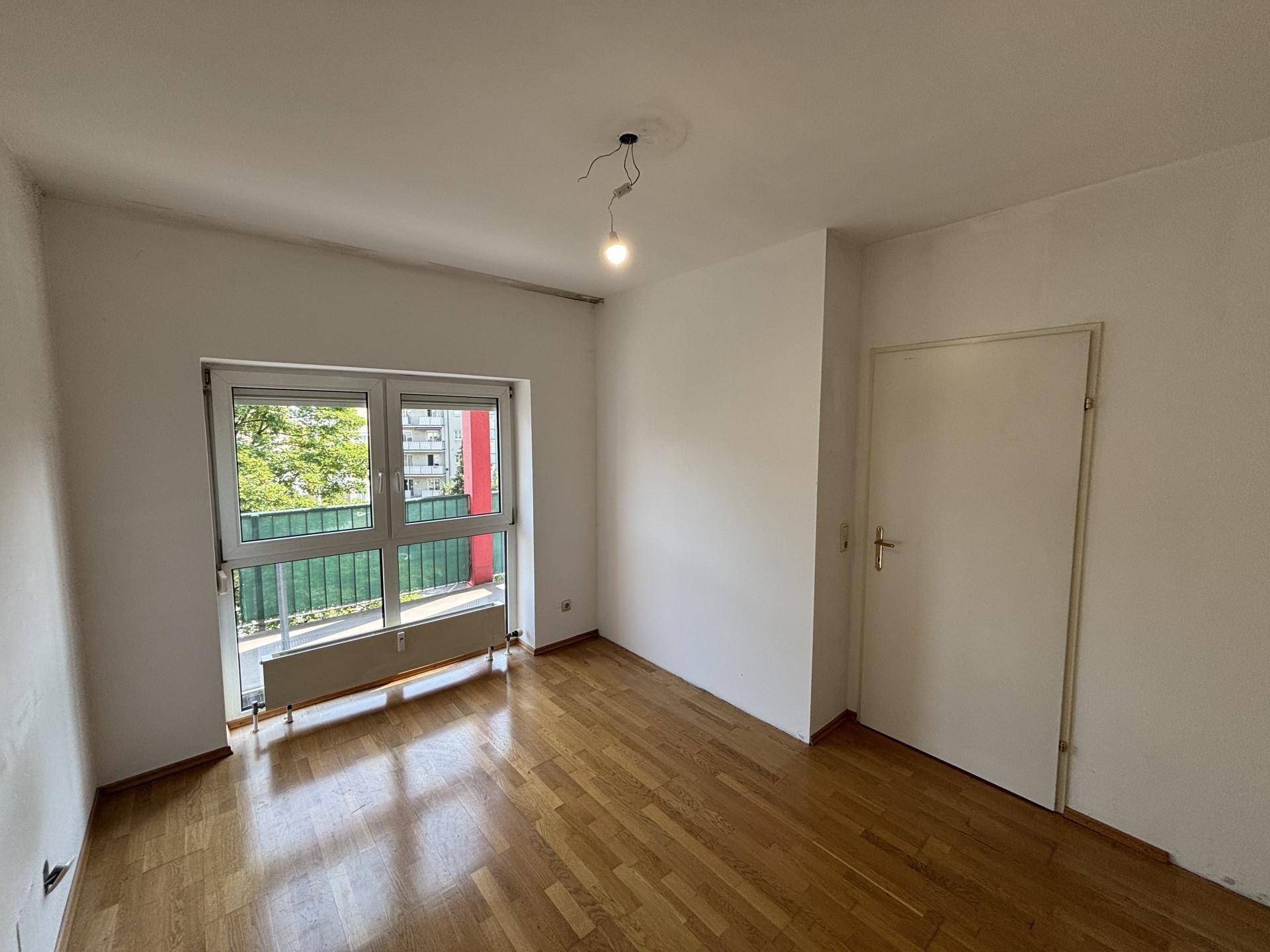 Helles Zimmer mit Holzboden und einem Fenster, das einen Blick auf eine rote Außenwand bietet.