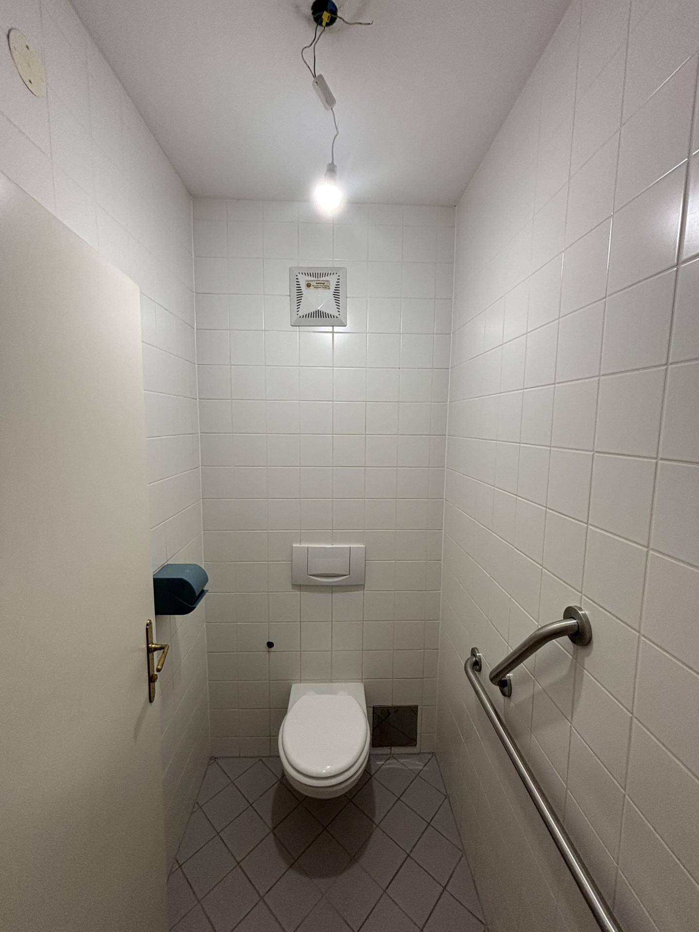 Kleines WC mit weißen Fliesen, wandhängender Toilette, Haltegriffen und einem Abluftventilator.