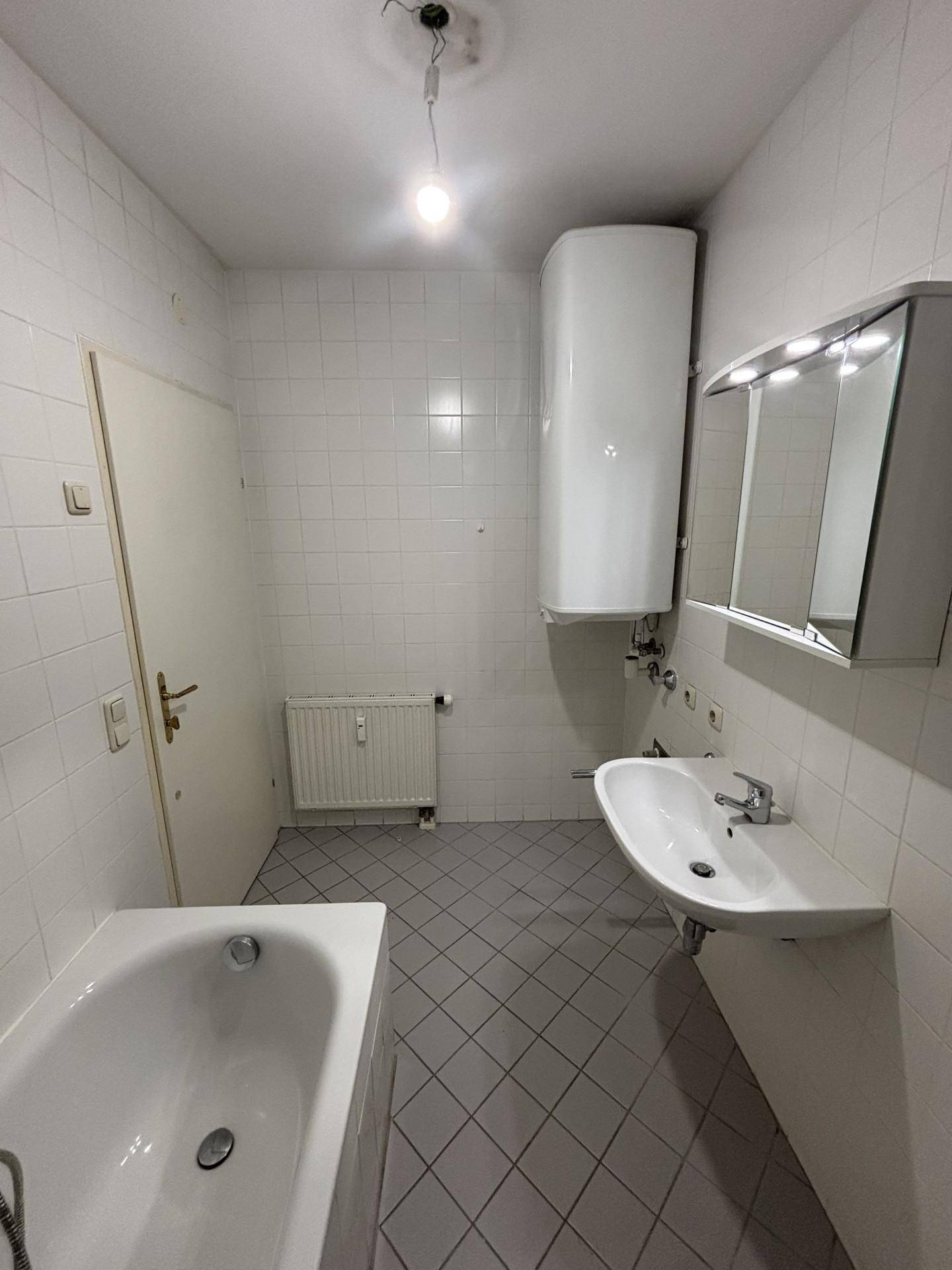 Badezimmer mit weißen Wandfliesen, grauen Bodenfliesen, Badewanne, Waschbecken und Warmwasserboiler.