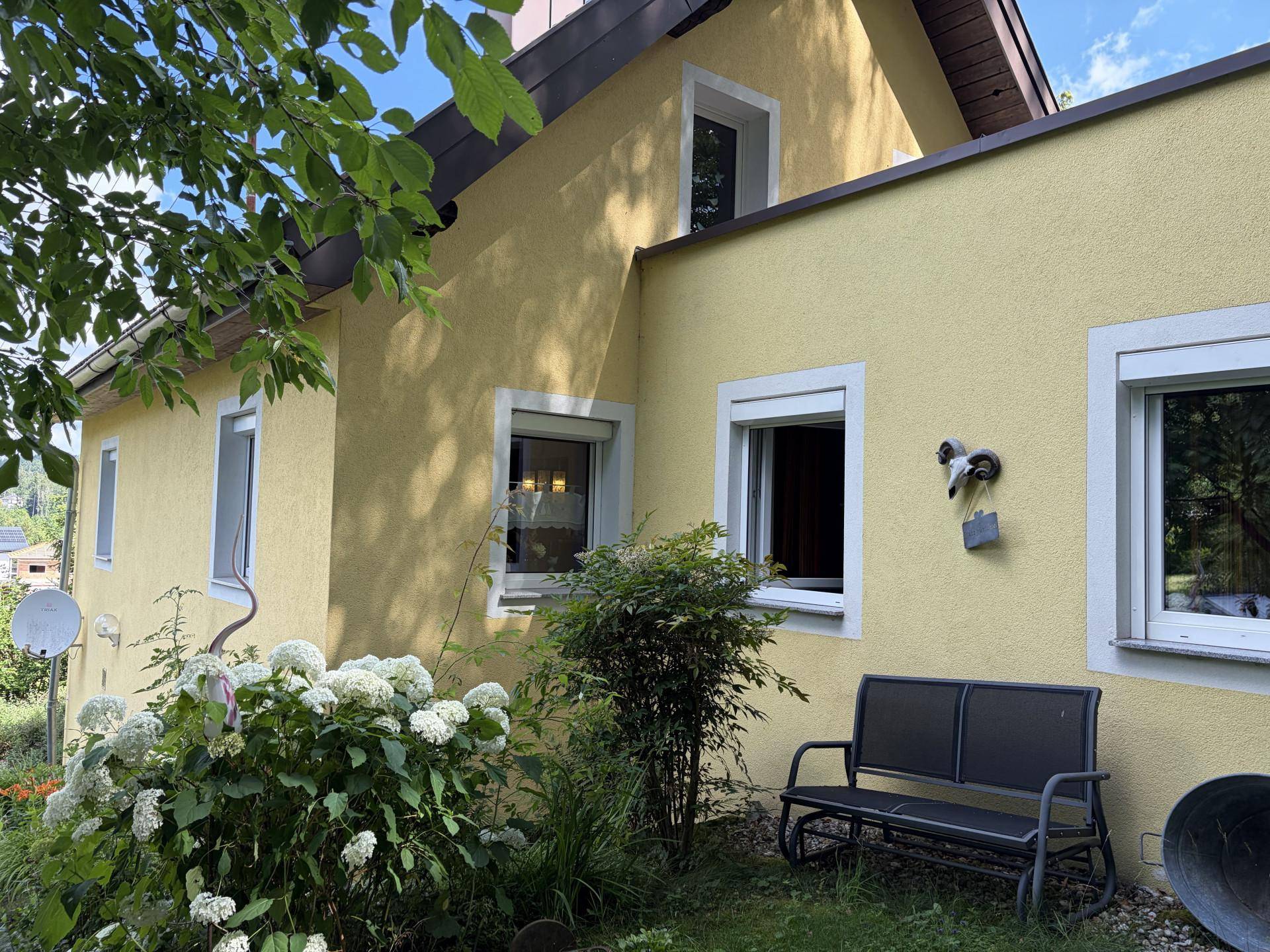 Seitenansicht des gelben Hauses mit kleinen Fenstern und einer einfachen Gartenbank im Grünen.