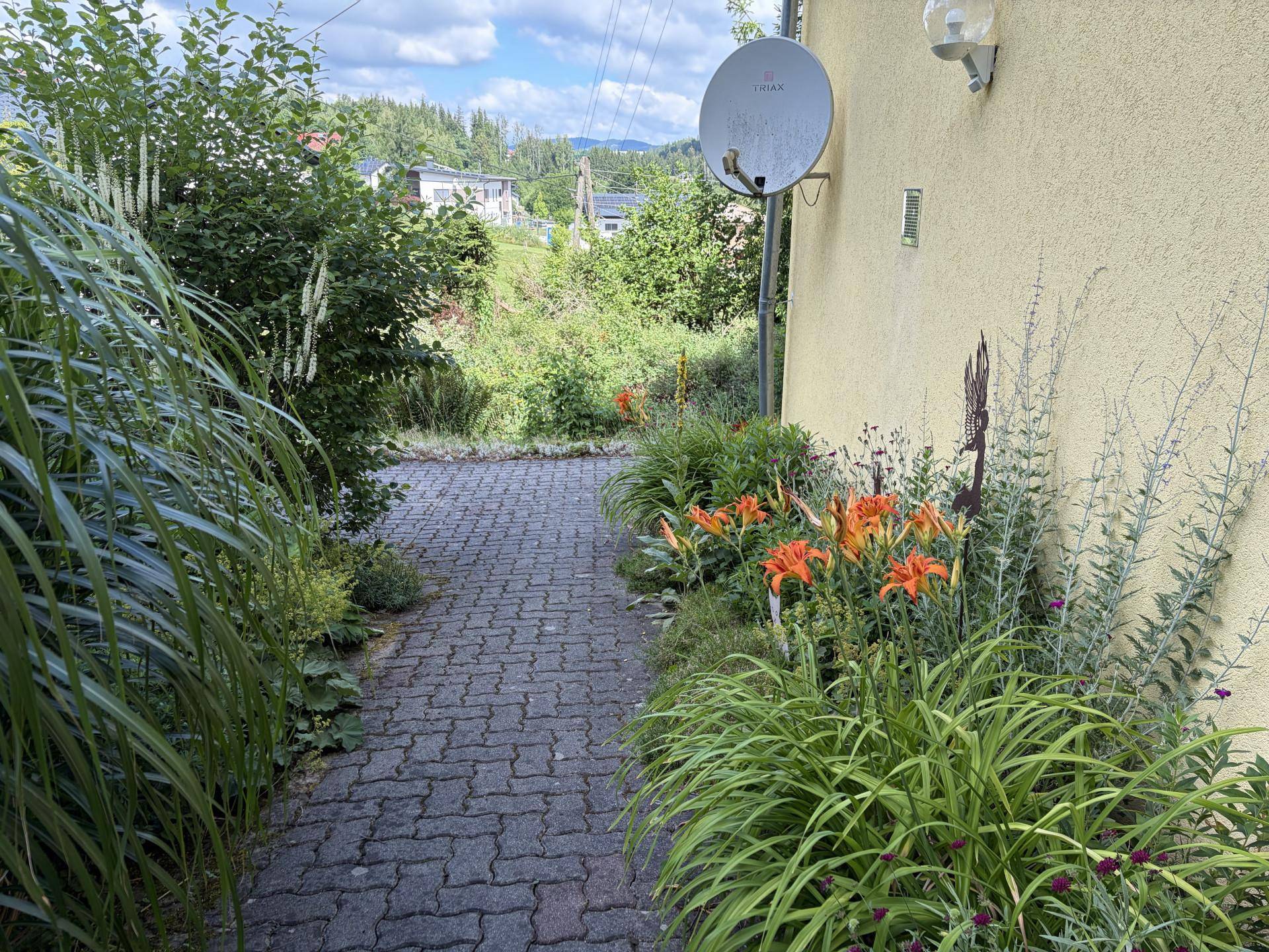 Gartenweg entlang des Hauses, gesäumt von Blumenbeeten und hohen Gräsern.