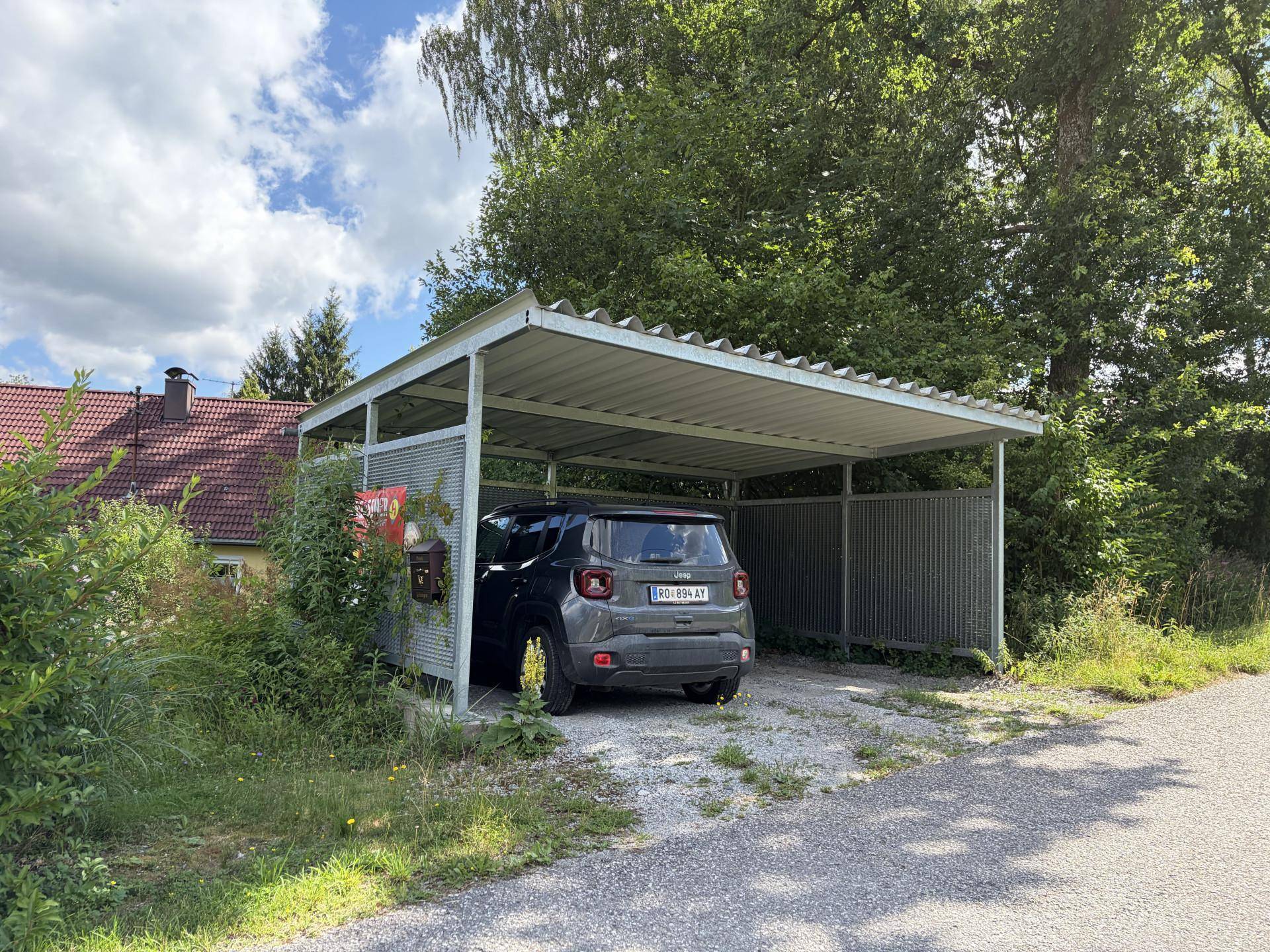 Überdachter Stellplatz für ein Fahrzeug, umgeben von Bäumen und Sträuchern.