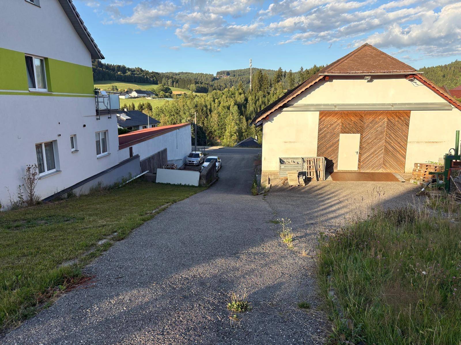 Blick auf das Gebäude und die Zufahrt mit ländlicher Umgebung im Hintergrund. Grüne Hügel sind sichtbar.
