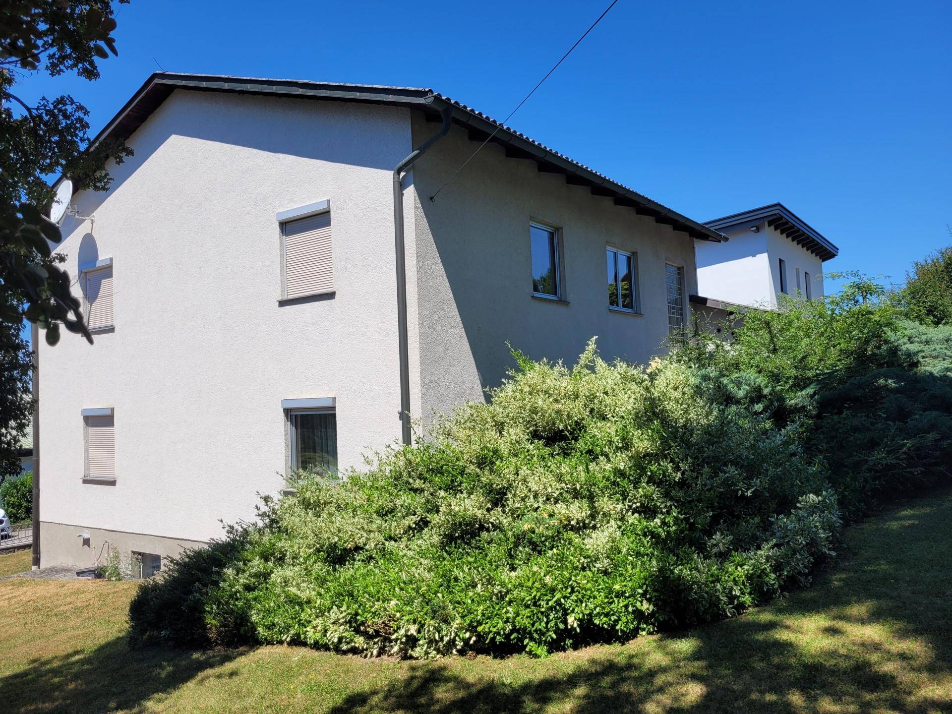 Helle Fassade eines Einfamilienhauses mit Rollläden und umgebender Bepflanzung unter blauem Himmel.