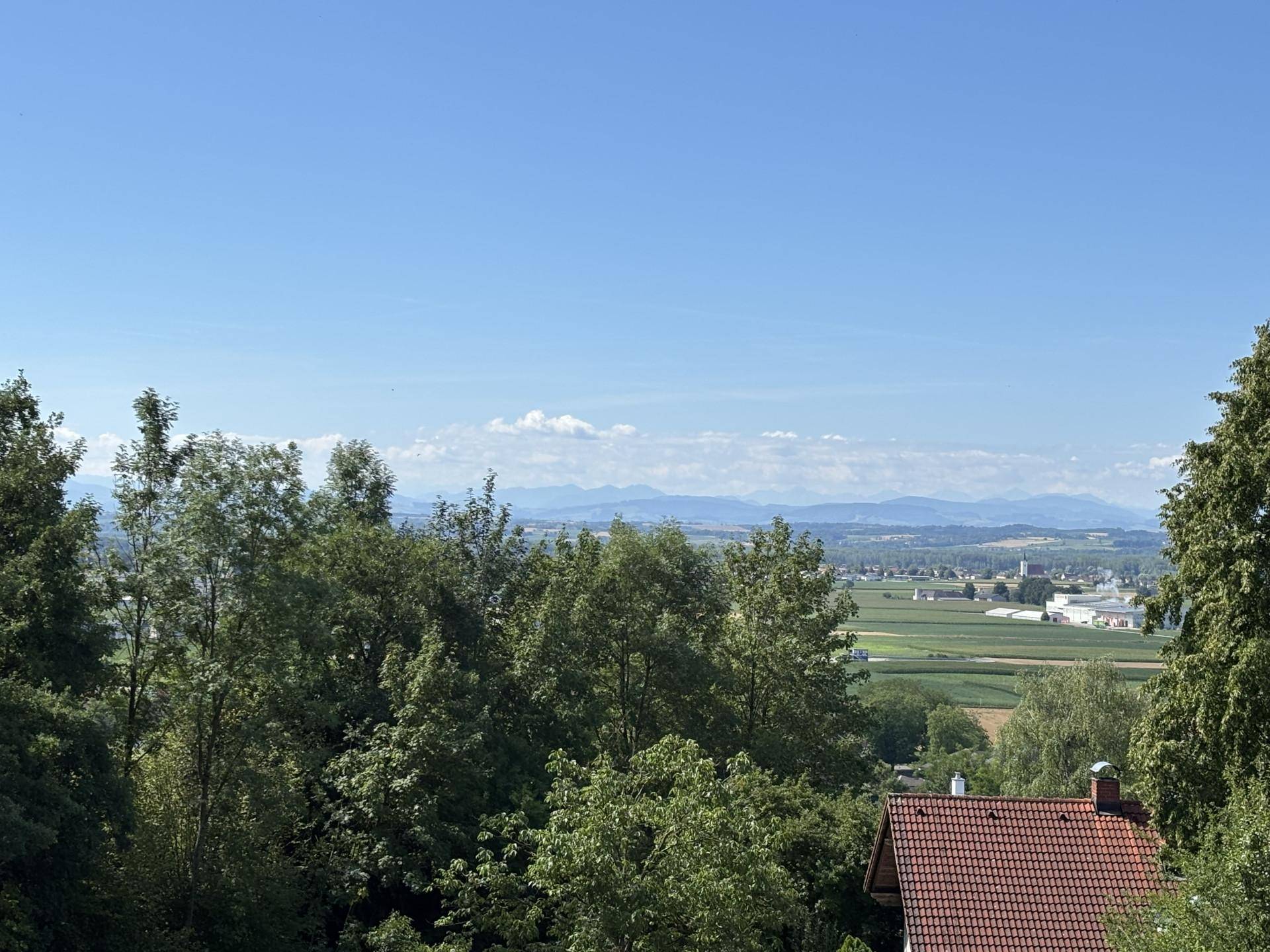 Weitläufiger Ausblick auf eine grüne Landschaft mit einem Haus und einer Bergkette am Horizont.