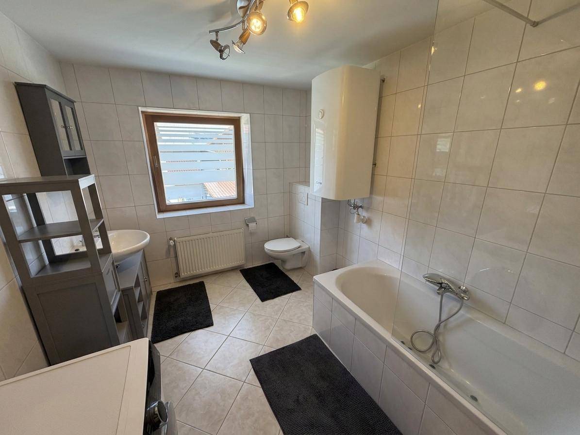 Helles Badezimmer mit Badewanne, WC, Waschbecken und Fenster, komplett mit weißen Fliesen ausgestattet.
