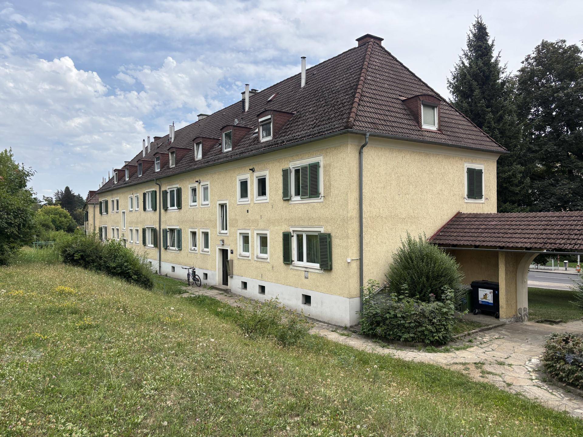 Gelbes Mehrfamilienhaus mit grünen Fensterläden und Satteldach, umgeben von einer Grünfläche.