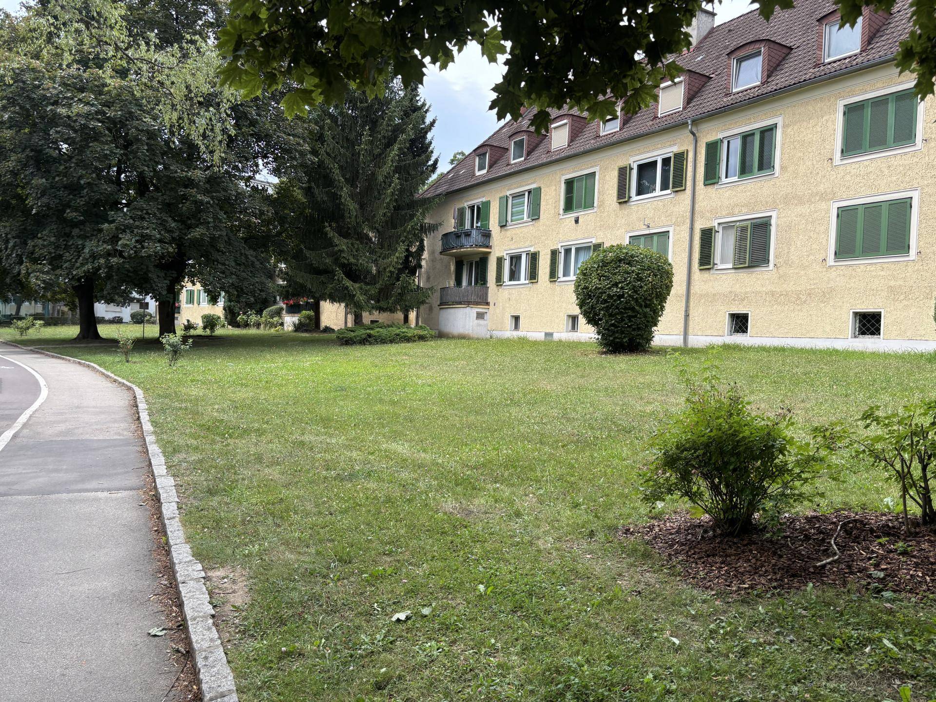 Seitenansicht des Gebäudes mit gepflegter Rasenfläche, hohen Bäumen und einem Gehweg.