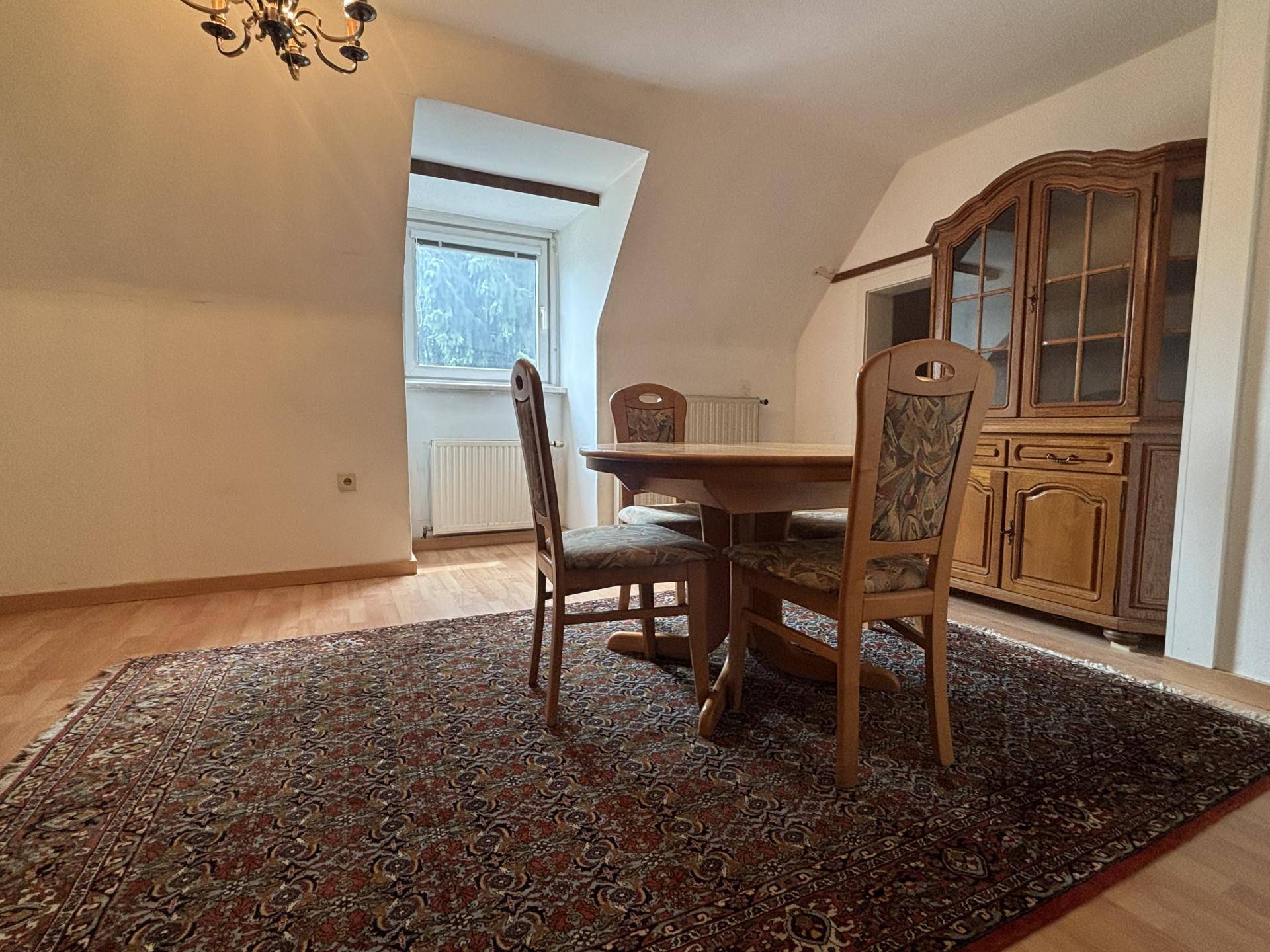 Heller Essbereich mit Holztisch, Stühlen und einem traditionellen Vitrinenschrank unter der Dachschräge.