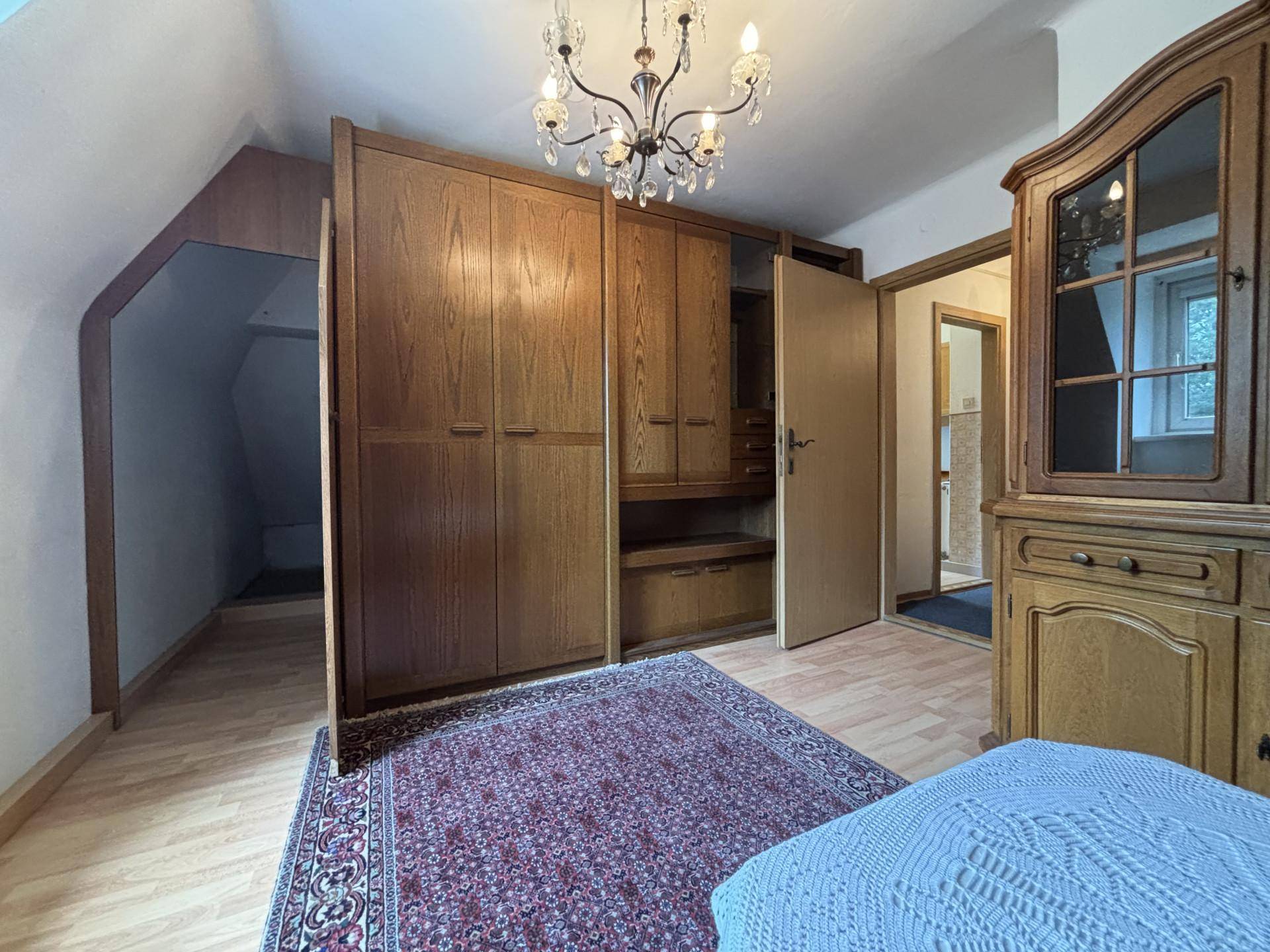 Schlafzimmer mit großem Holzschrank, Kronleuchter und Zugang zu einem weiteren Raum unter der Dachschräge.