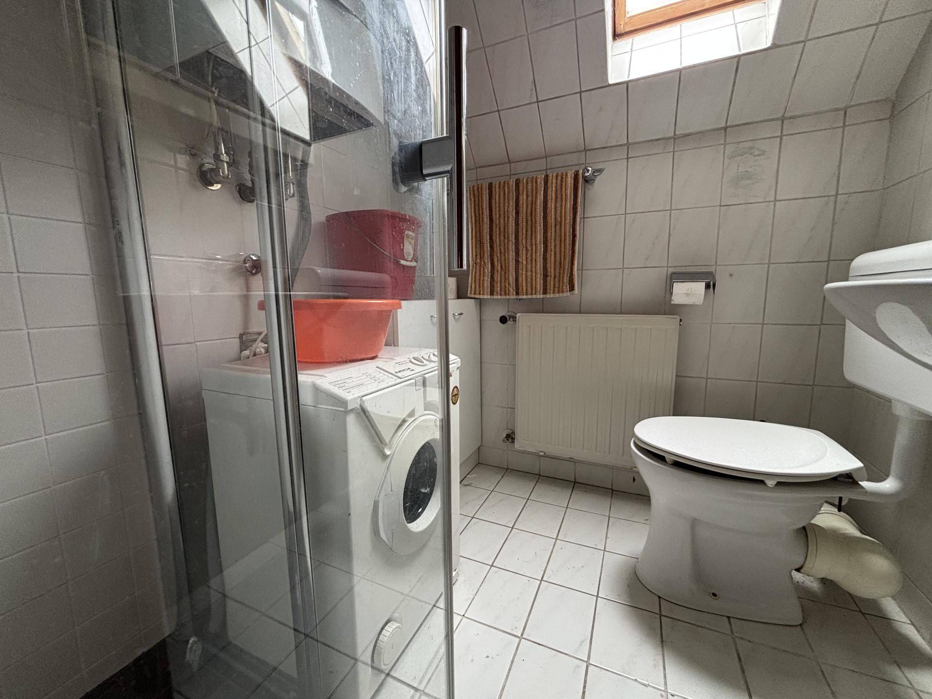 Badezimmer mit weißen Fliesen, Dusche, Toilette und Waschmaschine, ausgestattet mit einem Fenster.