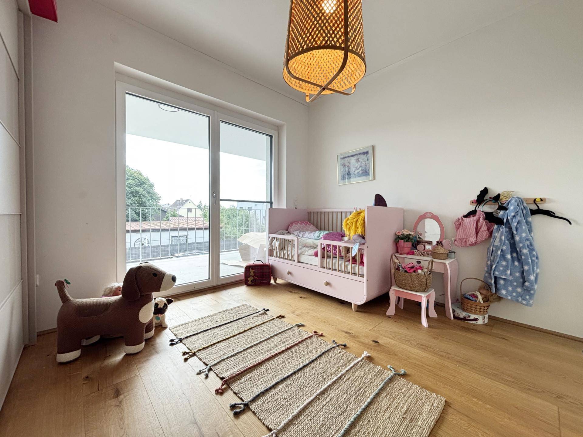 Kinderzimmer mit Babybett, Schminktisch und Zugang zum Balkon durch eine große Glastür.