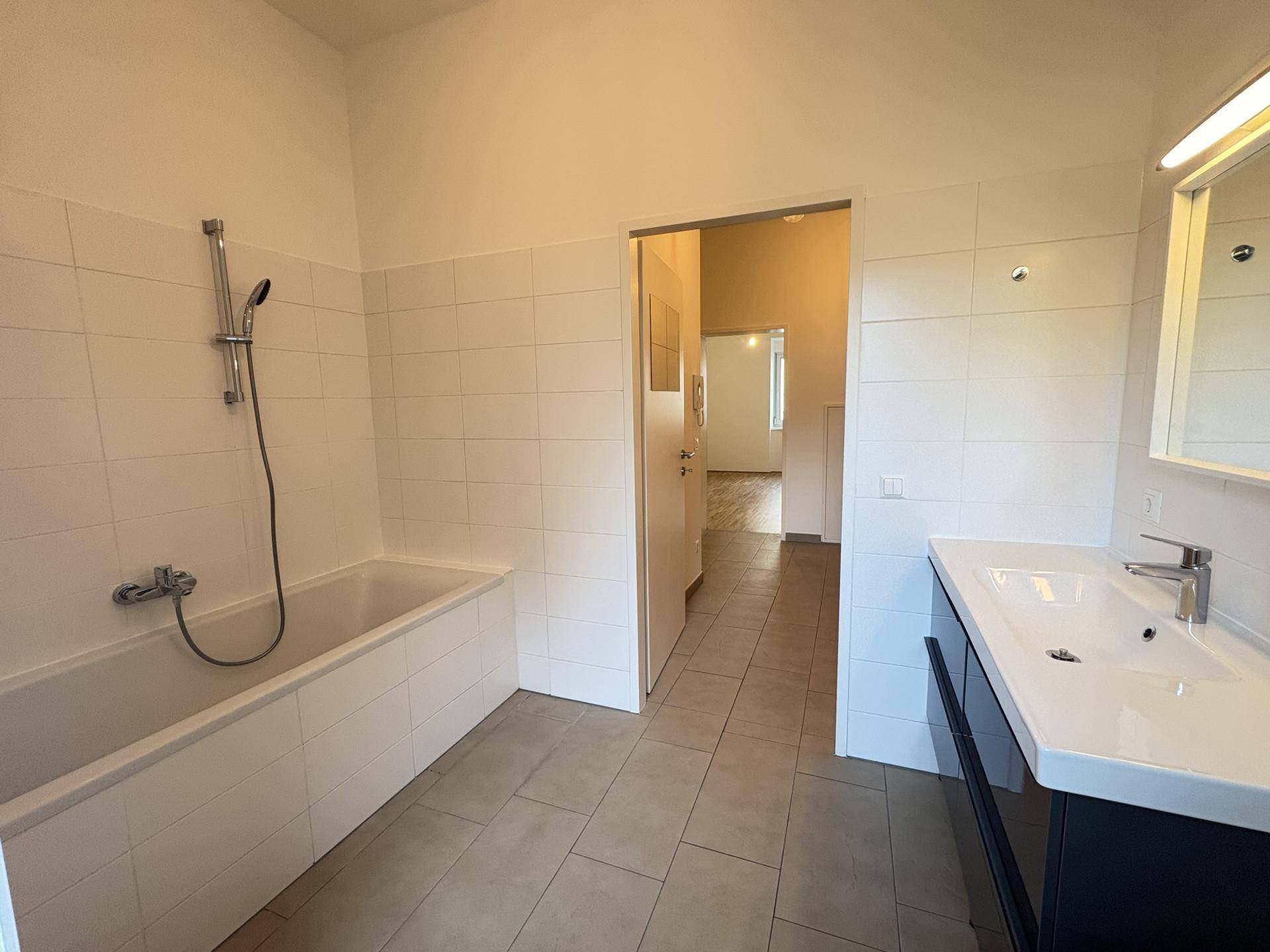 Helles Badezimmer mit Badewanne, Duscharmatur, Einzelwaschbecken und hellen Fliesen.