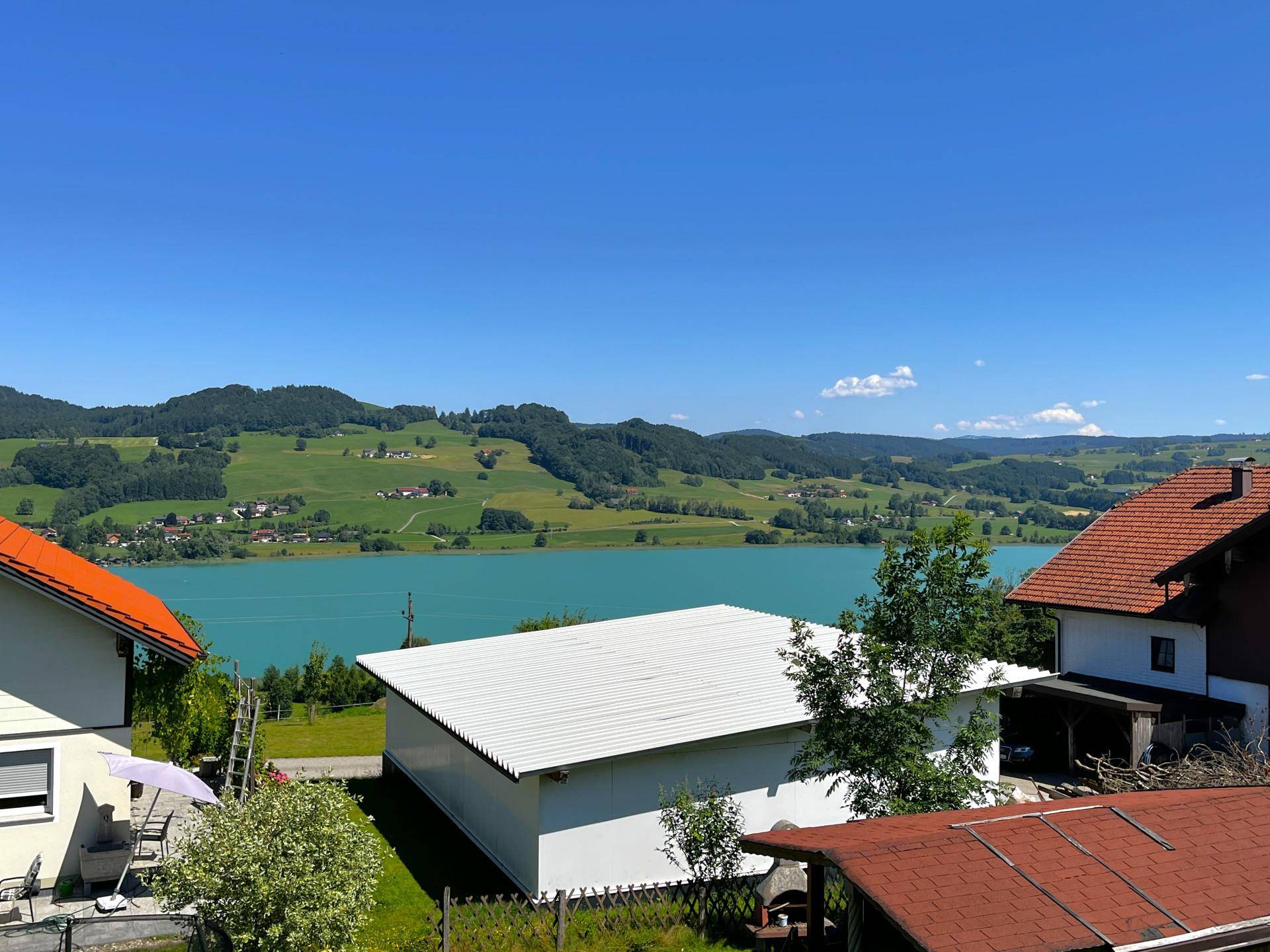 Panoramablick auf den See und die umliegende grüne Hügellandschaft an einem klaren, sonnigen Tag.