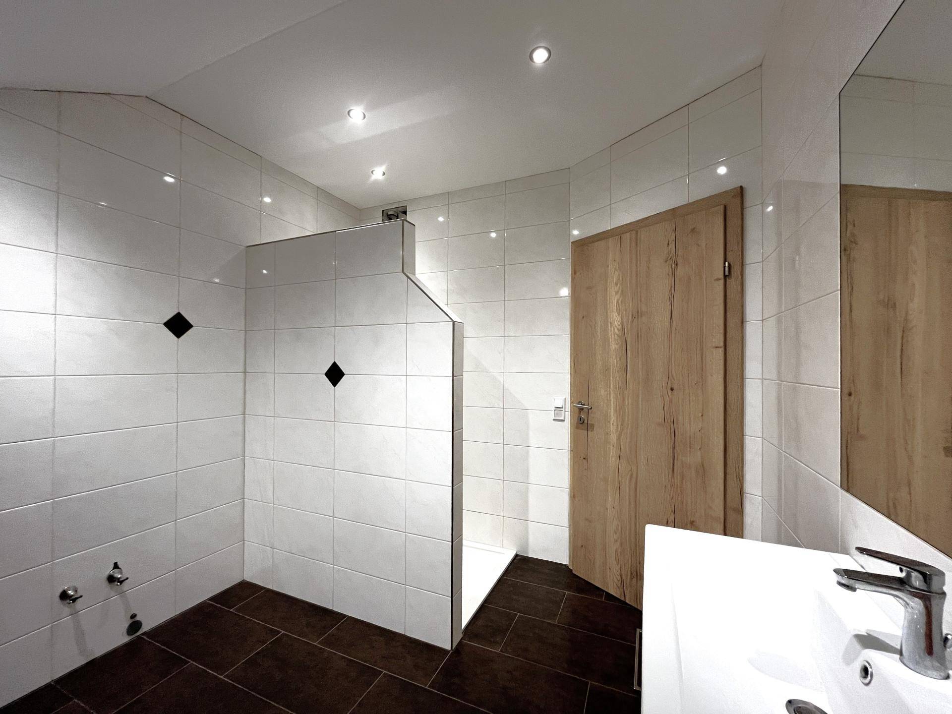 Geräumiges Badezimmer mit weißen Fliesen und dunklem Boden, vorbereitet für eine Dusche.